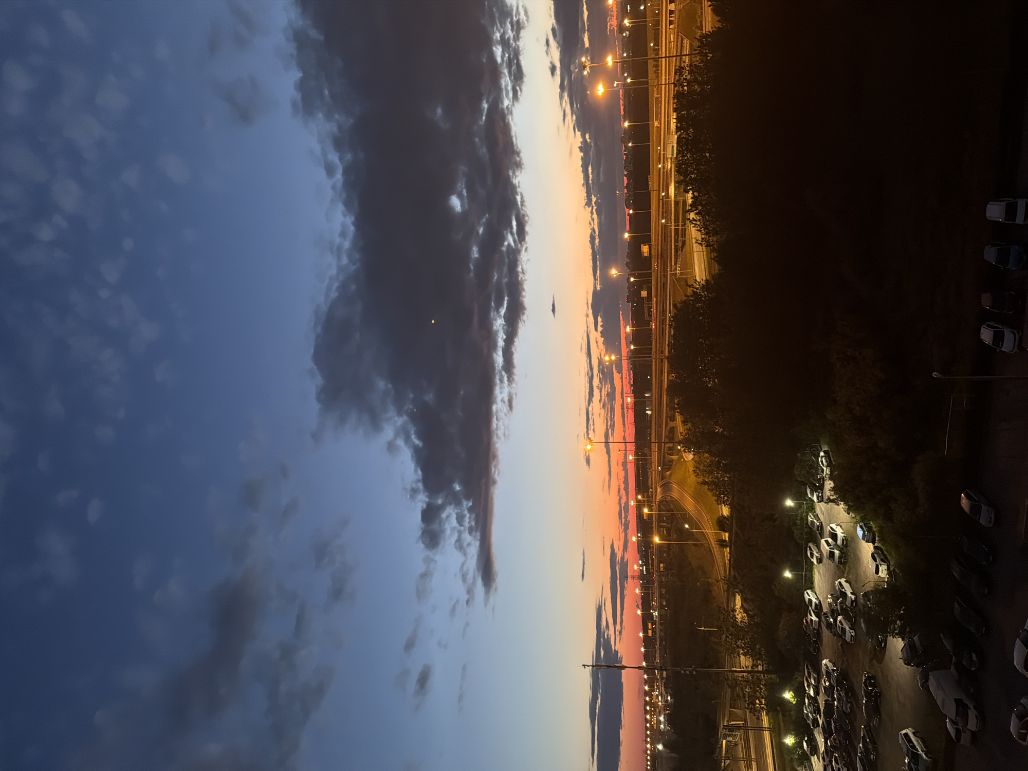 The image shows a twilight view of a city, possibly an airport or industrial area, with a parking lot in the foreground. The sky is a deep blue with streaks of orange and red near the horizon, indicating sunset. Dark clouds are scattered across the sky. Numerous streetlights illuminate a highway or elevated road system in the mid-ground. The foreground features a large, mostly empty parking lot filled with cars. The lighting suggests it is dusk or early evening. The scene is devoid of people and no specific actions or interactions are depicted. The environment appears to be an urban or suburban setting, characterized by infrastructure like roads and lighting.