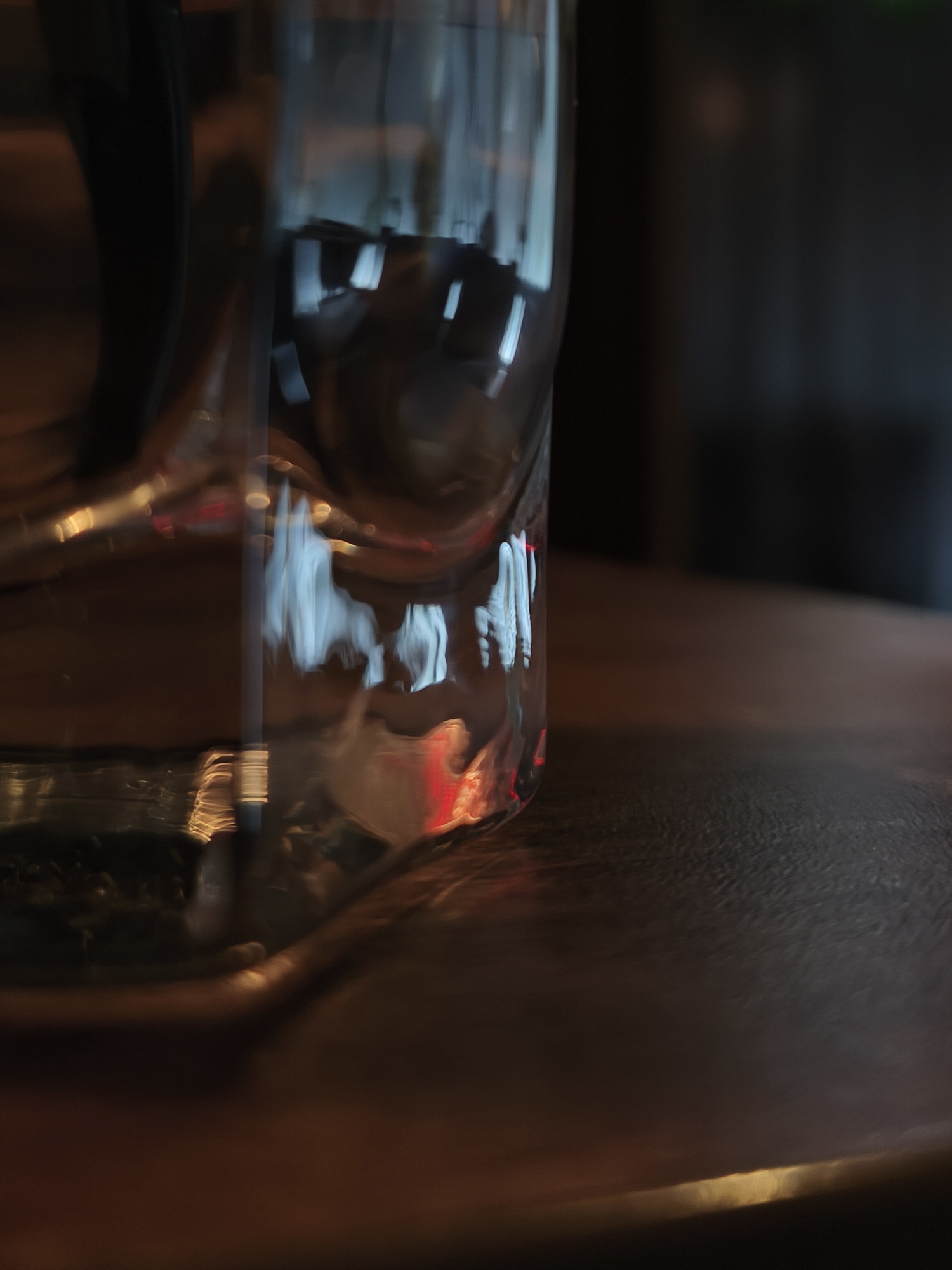 A close-up shot focuses on a clear glass, likely a drinking glass, standing on what appears to be a dark, possibly wooden, surface. The glass distorts the background, creating abstract light reflections that contain both red and white hues. Part of a black object is visible to the left. The background is blurred, suggesting an interior setting with a dark color palette. There is insufficient information to precisely identify this specific location in the city of Chaikovsky, Russia.