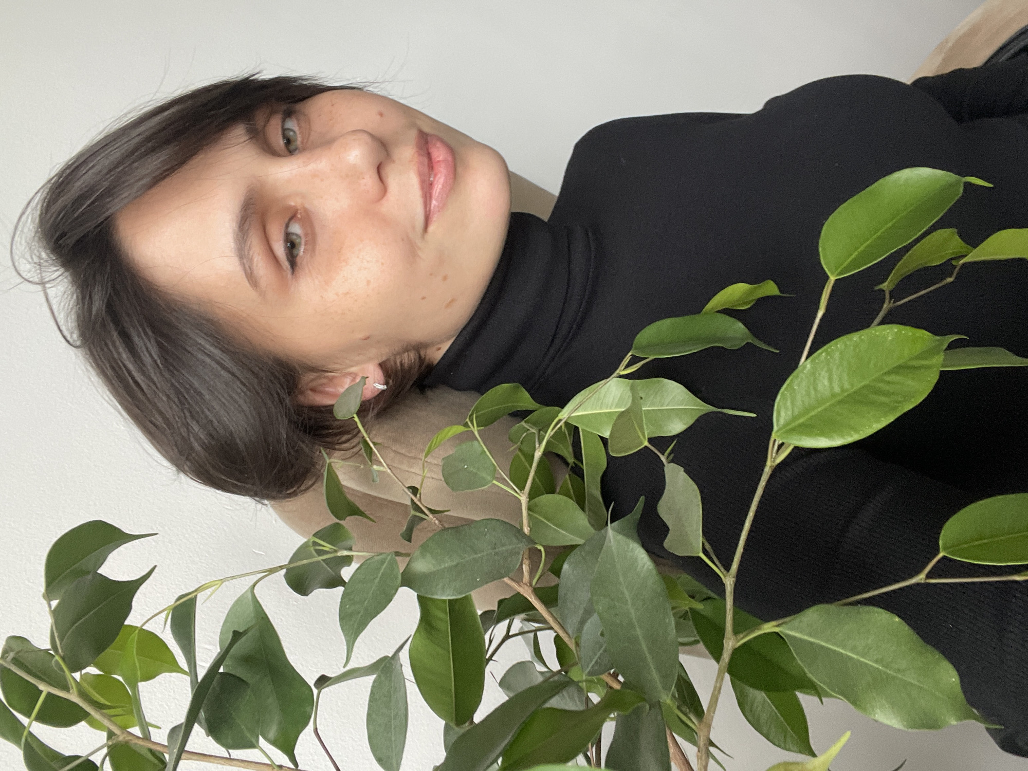 A close-up, indoor portrait features a young woman, likely taken in Izhevsk, Russia. The image is oriented with the woman's head tilted to the right, suggesting she is lying down or photographed from an elevated angle. She has short, dark brown hair, clear green eyes, and a scattering of freckles across her face. Her expression is serene with a slight, gentle smile. She is wearing a black turtleneck sweater, and a small, silver-toned earring is visible in her right ear. Her head rests against a light-colored cushion or furniture piece, set against a plain, light wall background.
Prominently featured in the foreground and partially overlapping the woman's body is a lush green houseplant. It has numerous oval-shaped leaves and slender branches. The lighting is bright and even, indicating a daytime setting indoors. No text is visible in the image.