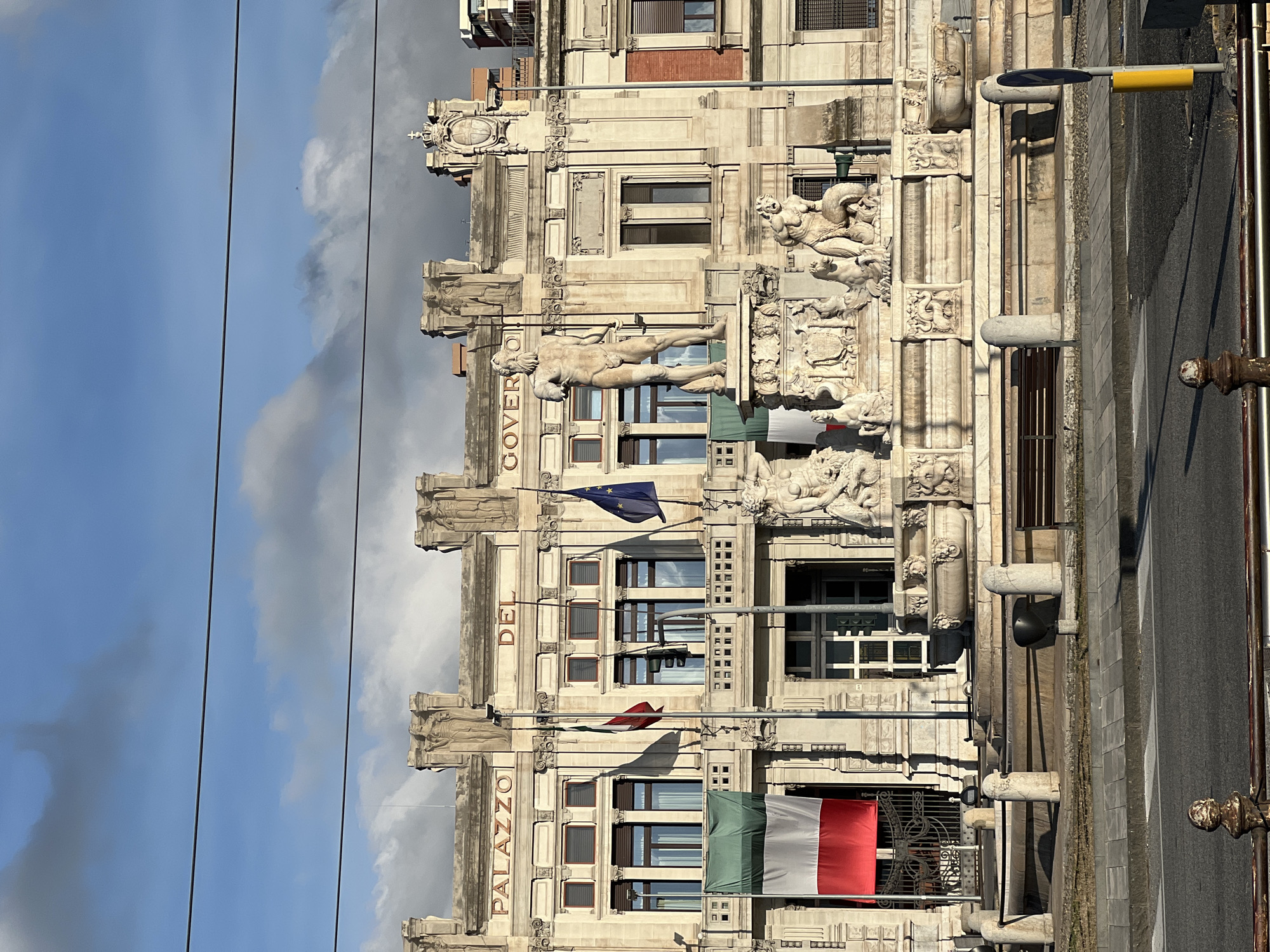The image presents an urban scene dominated by the Palazzo del Governo and the Fountain of Neptune in Messina, Italy. The Palazzo del Governo is a multi-story, light-colored stone building with neoclassical-style architecture, featuring "PALAZZO DEL GOVERNO" inscribed in raised letters above its upper windows. The building's facade is adorned with decorative elements including pilasters, balconies, and sculpted figures, with a prominent crest visible on the upper right side.
Three flags are displayed: two Italian flags (green, white, and red vertical stripes) – one flying from a pole on the left and another draped vertically from a balcony on the far left – and a European Union flag (blue with twelve yellow stars) flying from a pole on the right. A traffic light fixture is visible between the two flagpoles.
In front of the building stands the elaborate Fountain of Neptune, constructed from light-colored stone. A large, nude male statue, holding a trident-like staff, dominates the center of the fountain, atop a multi-tiered base. The base features numerous sculpted figures, including mythological sea creatures, lions, and other decorative carvings, with water basins incorporated into its design.
The foreground shows a paved street with a white-striped pedestrian crosswalk. A wide sidewalk separates the street from the fountain, featuring stone bollards, a brown bench, and a low metal barrier. Sunlight illuminates the scene from the right, casting distinct shadows across the fountain and street. The sky above is partly cloudy, with areas of clear blue visible between large, grey cumulus clouds. No living individuals are present in the immediate vicinity of the main subjects.