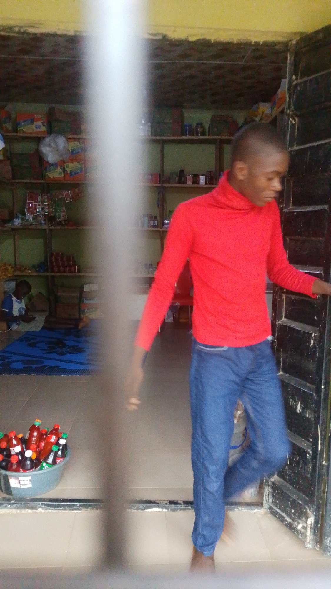 A young Black male wearing a red turtleneck shirt and blue jeans walks through a doorway. He is barefoot. In the background, shelves are stocked with various items, including food products and bottles. To the left, another young Black male sits on a blue mat, looking at a phone. A tub of bottled drinks is visible in the foreground. The scene appears to be inside a small shop or market.