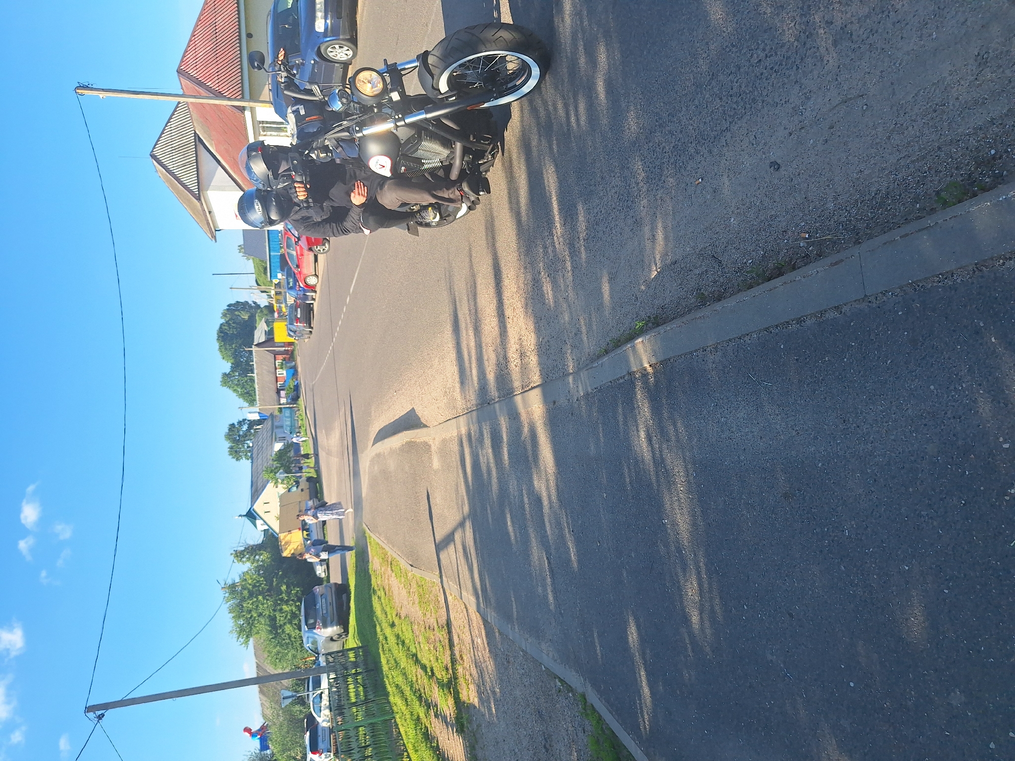 The image captures a street scene in Блеўчыцкі сельскі Савет, Belarus. In the foreground, two individuals, a man and a woman, are riding a motorcycle. Both are wearing helmets. The man is driving the motorcycle, and the woman is seated behind him. They are dressed in casual clothing. The motorcycle is dark in color and appears to be a cruiser style. The road is paved and has a sidewalk on the left side. In the background, there are several houses with red and white roofs, a few parked cars, trees, and a utility pole with wires. The sky is clear and blue, indicating a sunny day. The overall environment suggests a rural or suburban setting within Belarus.