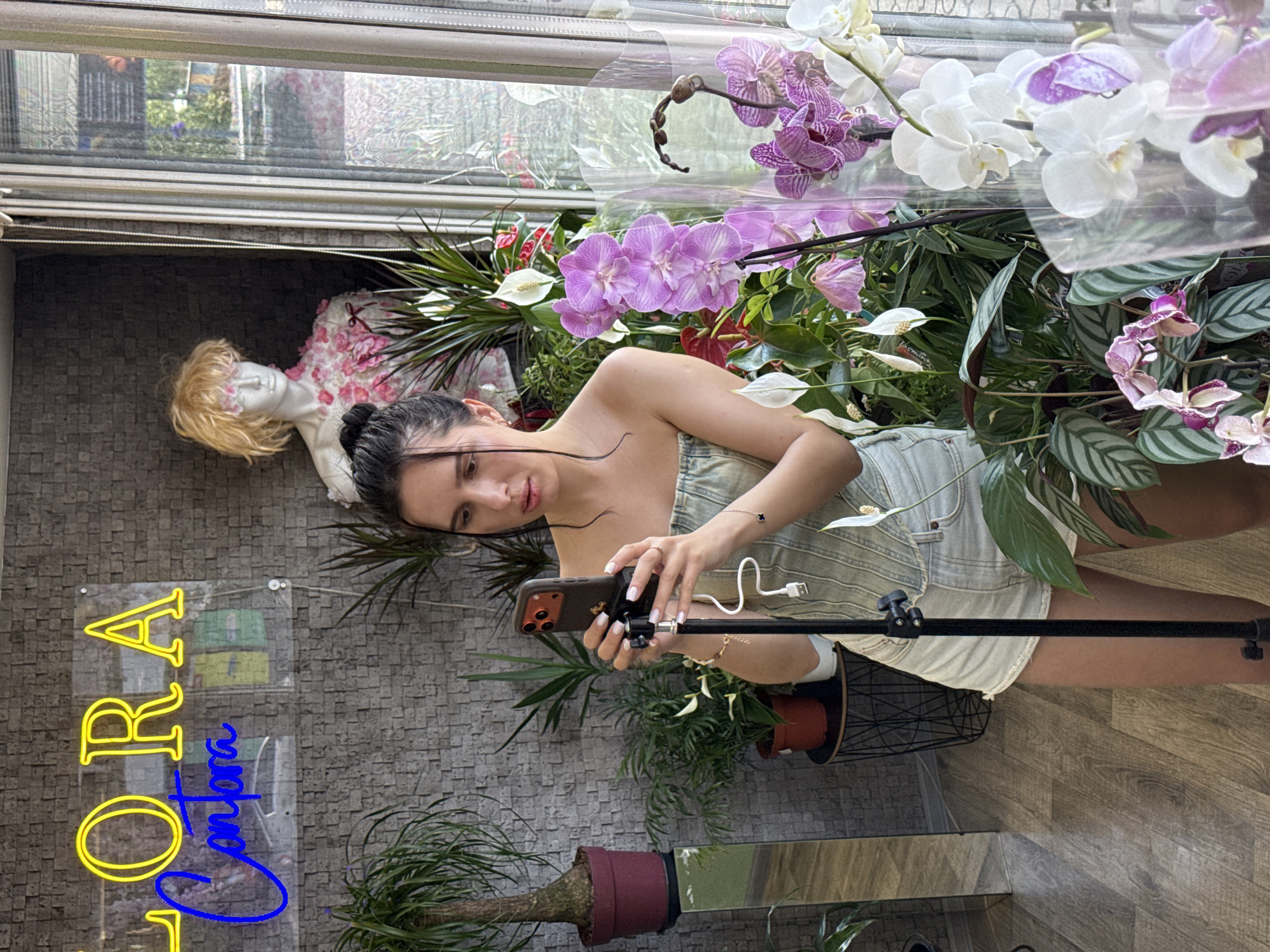 A young woman with dark hair tied back is holding a smartphone on a tripod in front of her, in what appears to be a flower shop. She is wearing a light-colored, strapless top and denim shorts. The background is filled with various potted plants and orchids, with a mannequin adorned with pink flowers visible behind her. A neon sign reading "FLORA Comforn" is mounted on a grey brick wall. A window is to the right of the scene, offering a glimpse of the outside. The floor is made of wooden planks.