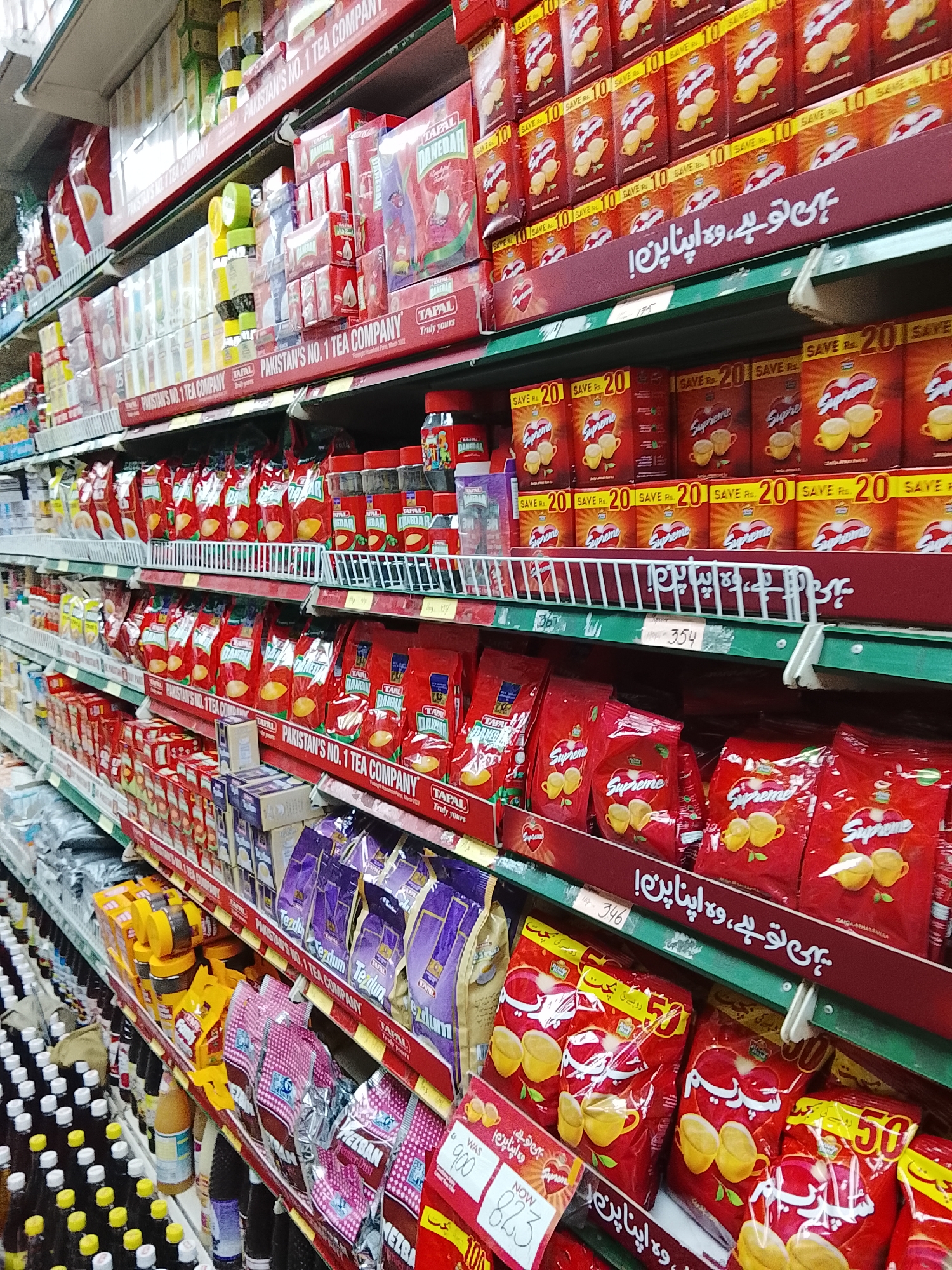 The image shows a shelf in a supermarket in Gulgasht Colony, Pakistan. The shelf is stocked with various tea products, mostly from the brand Tapal, which is advertised as "Pakistan's No. 1 Tea Company." Different varieties are visible, including Tapal Danedar and Tapal Supreme. Price tags and promotional signs like "SAVE Rs. 20" and "SAVE Rs. 10" are present. The shelf's price tag lists "WAS 900, NOW 823." The shelving is lined with dark brown trim and green accents. A few drinks are present on the lowest shelf. The scene suggests a typical grocery shopping environment.
