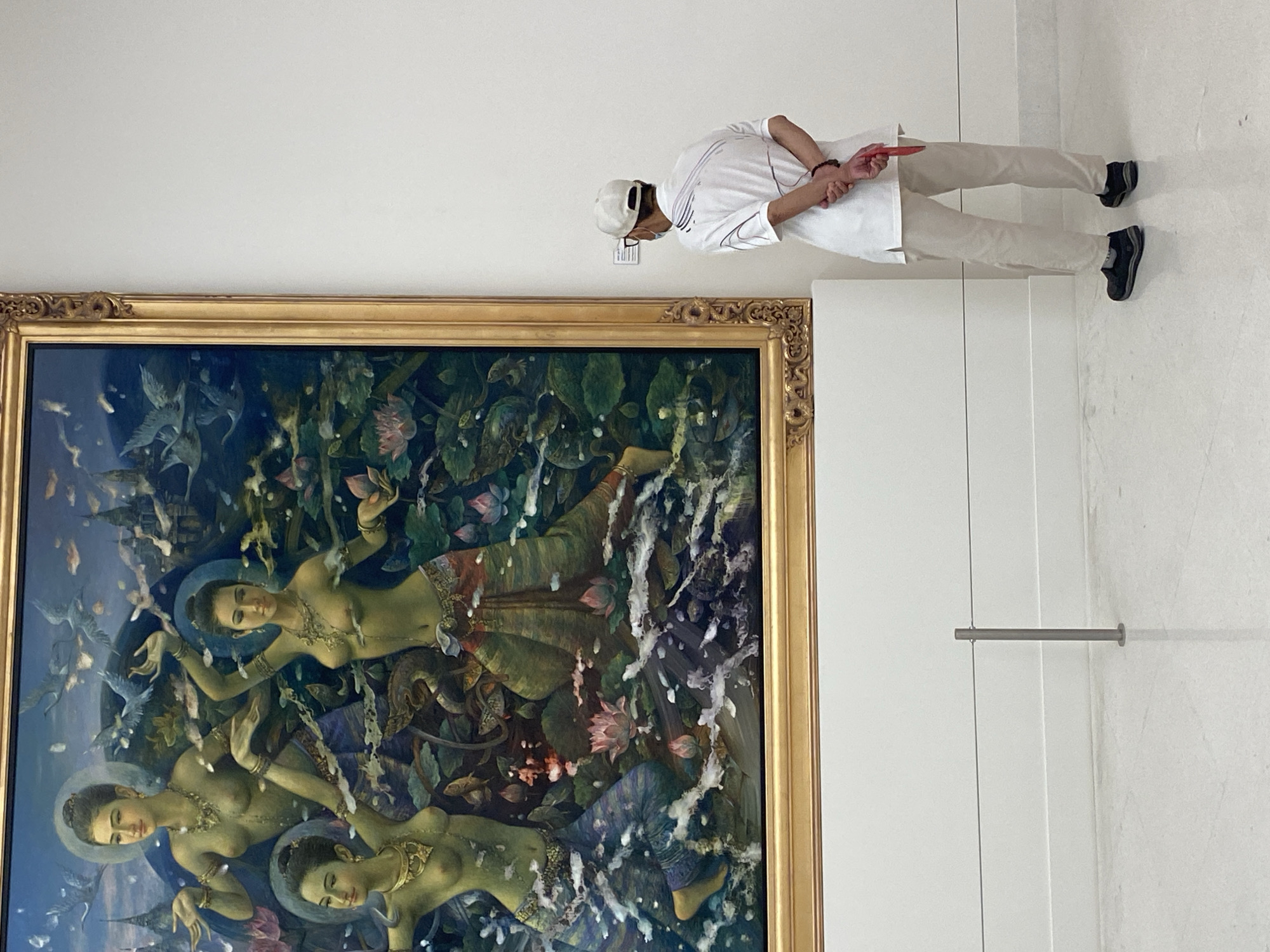 A man in a white shirt, light pants, and a white cap stands in an art gallery, viewing a large painting. The painting, framed in gold, depicts three partially clad female figures in a lush aquatic setting with birds, lotus flowers, and vegetation. The setting of the image is implied to be Bangkok, Thailand.