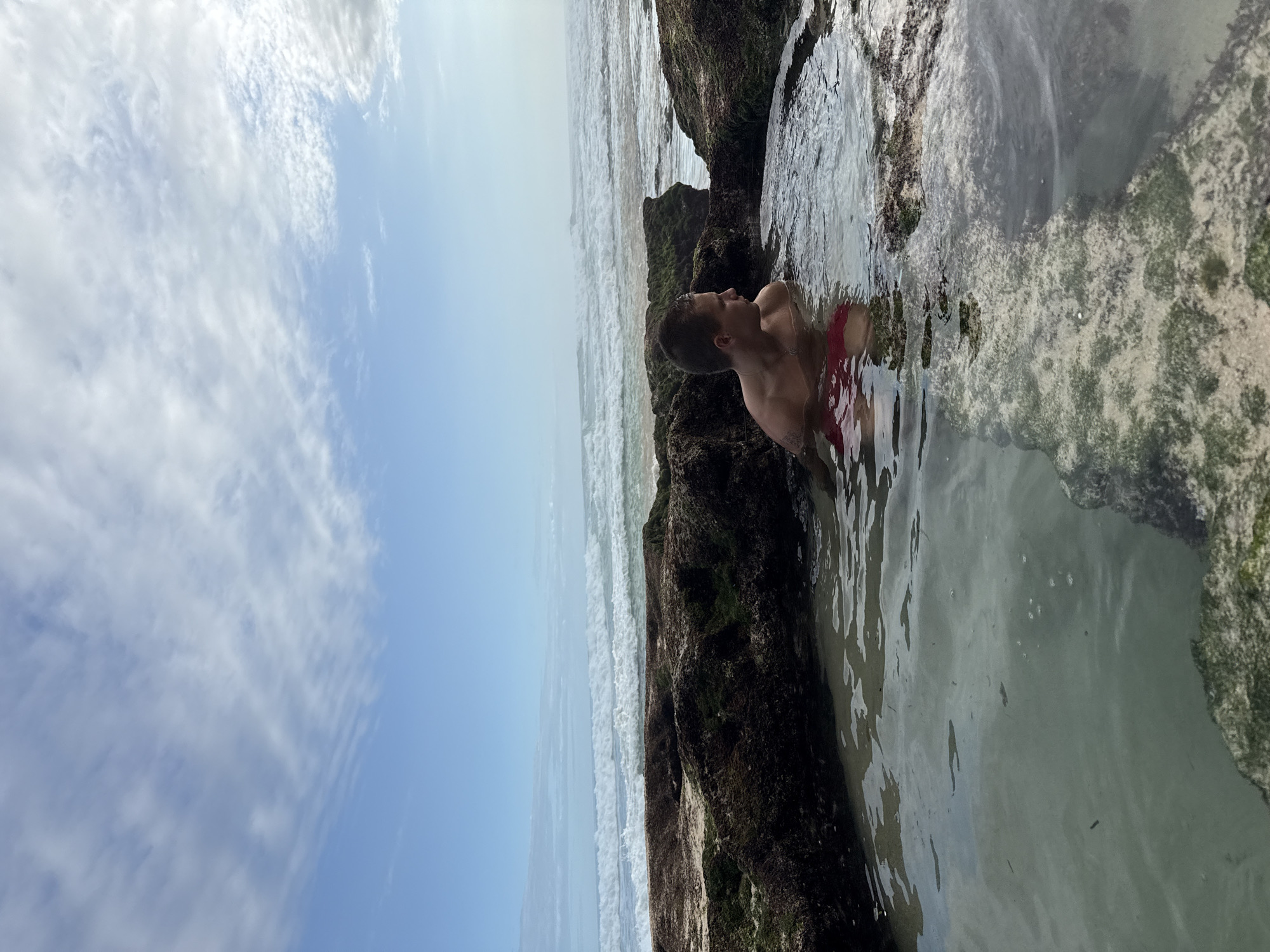 A medium-shot shows a male person in red swimming shorts in a shallow pool of water formed by dark-colored rocks covered with moss and algae. The ocean is in the background. The image was taken during daylight in Kuta Selatan, Indonesia. The sky above features a mix of blue and white clouds. The pool is shallow enough to see the sand at the bottom.