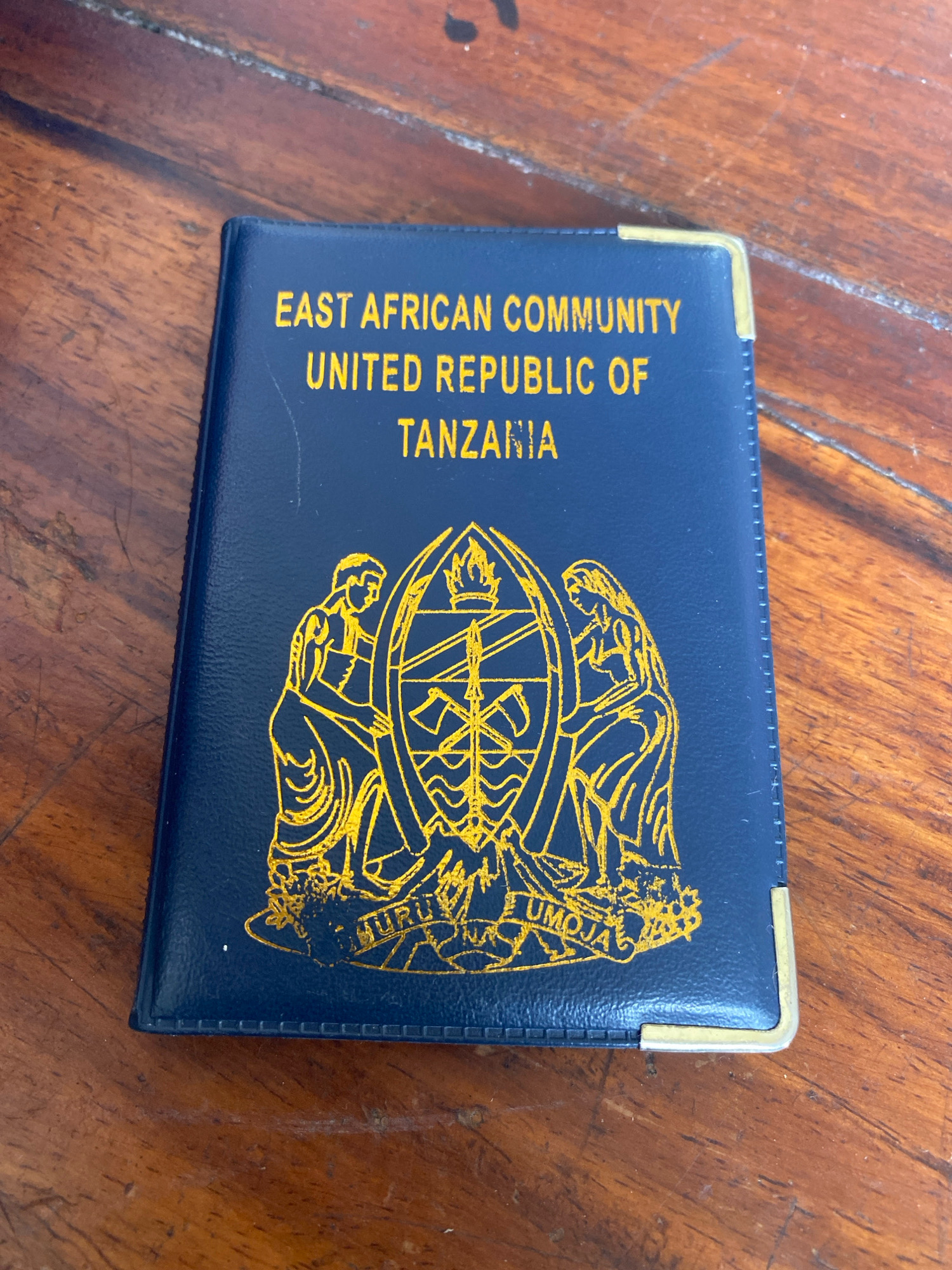 A dark blue passport, adorned with gold lettering and the national emblem of Tanzania, rests on a wooden surface. The passport, embossed with "EAST AFRICAN COMMUNITY" and "UNITED REPUBLIC OF TANZANIA", is positioned at an angle, with its bottom left corner slightly closer to the viewer. The emblem features two figures, one on each side of a shield-like object, with the words "UHURU" and "UMOJA" inscribed below. The wooden surface appears to be a table with visible grain patterns and a slightly worn texture. The lighting suggests an indoor environment, possibly a home or office. The image provides no contextual information about the city of Bububu, Tanzania.
