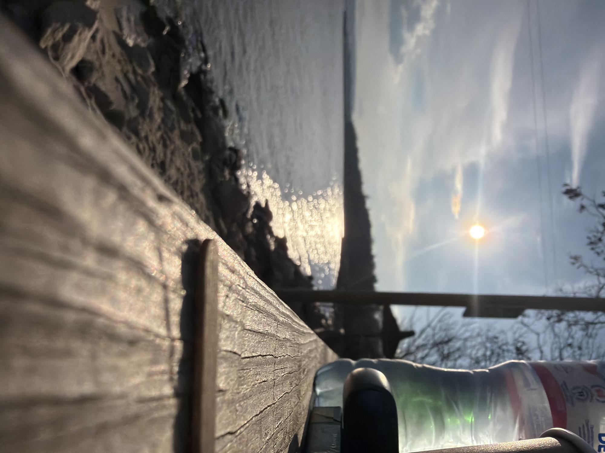 The image depicts a view over a body of water towards the horizon, presumably the Black Sea, likely taken from a location in Varna, Bulgaria. In the foreground, a weathered wooden railing extends along the left side of the frame. To the right of the railing, a water bottle is visible on what appears to be a horizontal surface, possibly a bench or wall. The sun is high in the sky, creating a bright glare and reflections on the water's surface. The sky is partly cloudy. Trees and other vegetation are visible on the right side of the frame, along the shoreline. The horizon is a long, faint line, suggesting a significant distance.