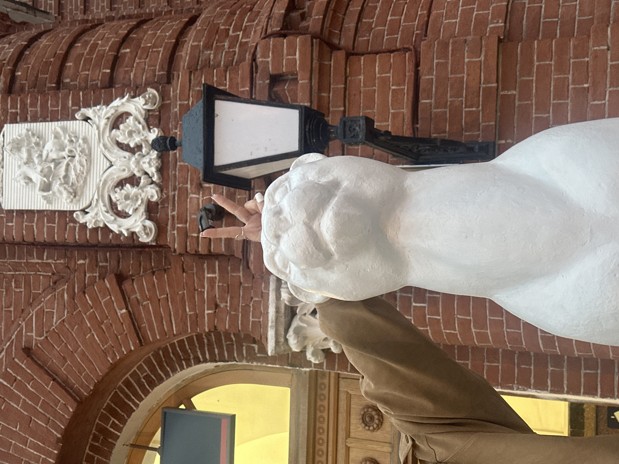This image captures a partial view of a person interacting with a white sculpture against a detailed red brick building in Kazan, Russia. The building exhibits an arched doorway or window on the left, framed by decorative light-colored wood featuring circular carved rosettes. A bright yellow light emanates from within the arched opening. Mounted on the red brick wall, above the sculpture, is a white plaster or stone relief depicting the coat of arms of Kazan, featuring a crowned winged dragon (Zilant) struck by a spear, set within an ornate, scrolled cartouche. To the right of the sculpture and below the coat of arms, a black, antique-style lamppost with a white opaque pane is affixed to the brickwork. The brick wall itself displays a curved, possibly turretted, section on the right side of the frame.
The central subject is a smooth, matte white sculpture of an animal bust, resembling a stylized feline such as a lion or panther, depicted with a prominent head, rounded ears, and a stout neck. The sculpture lacks detailed facial features. A person's right arm, wearing a light brown, long-sleeved garment, extends from the lower left of the frame. The person's right hand is positioned above the sculpture's head, forming a "peace" sign (V-sign) with the index and middle fingers extended upwards. A metallic ring is visible on the ring finger, and the fingernails are painted a light, possibly white, color.