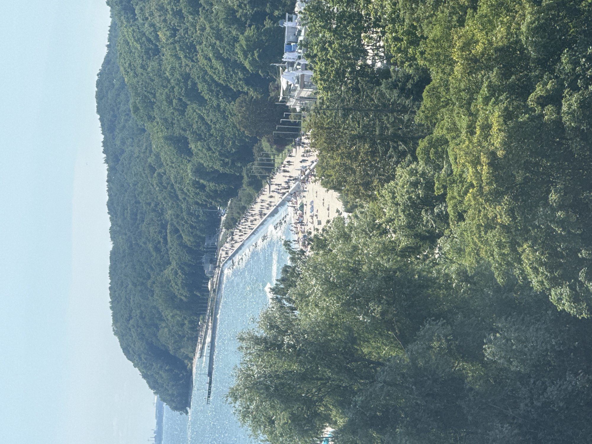 This is a high-angle, eye-level, outdoor, daytime, wide shot of the Gdynia coastline in Poland. The foreground is dominated by dense, green foliage of trees. Behind the trees, a long, paved promenade runs parallel to a sandy beach bordering a blue, choppy sea. Numerous people are visible walking along the promenade and sitting on the beach. A pier extends into the sea on the left side of the image. Buildings with white umbrellas and white structures line the promenade, suggesting a recreational or commercial area. In the distance, a forested hill rises from the coastline. The sky is clear and light blue.