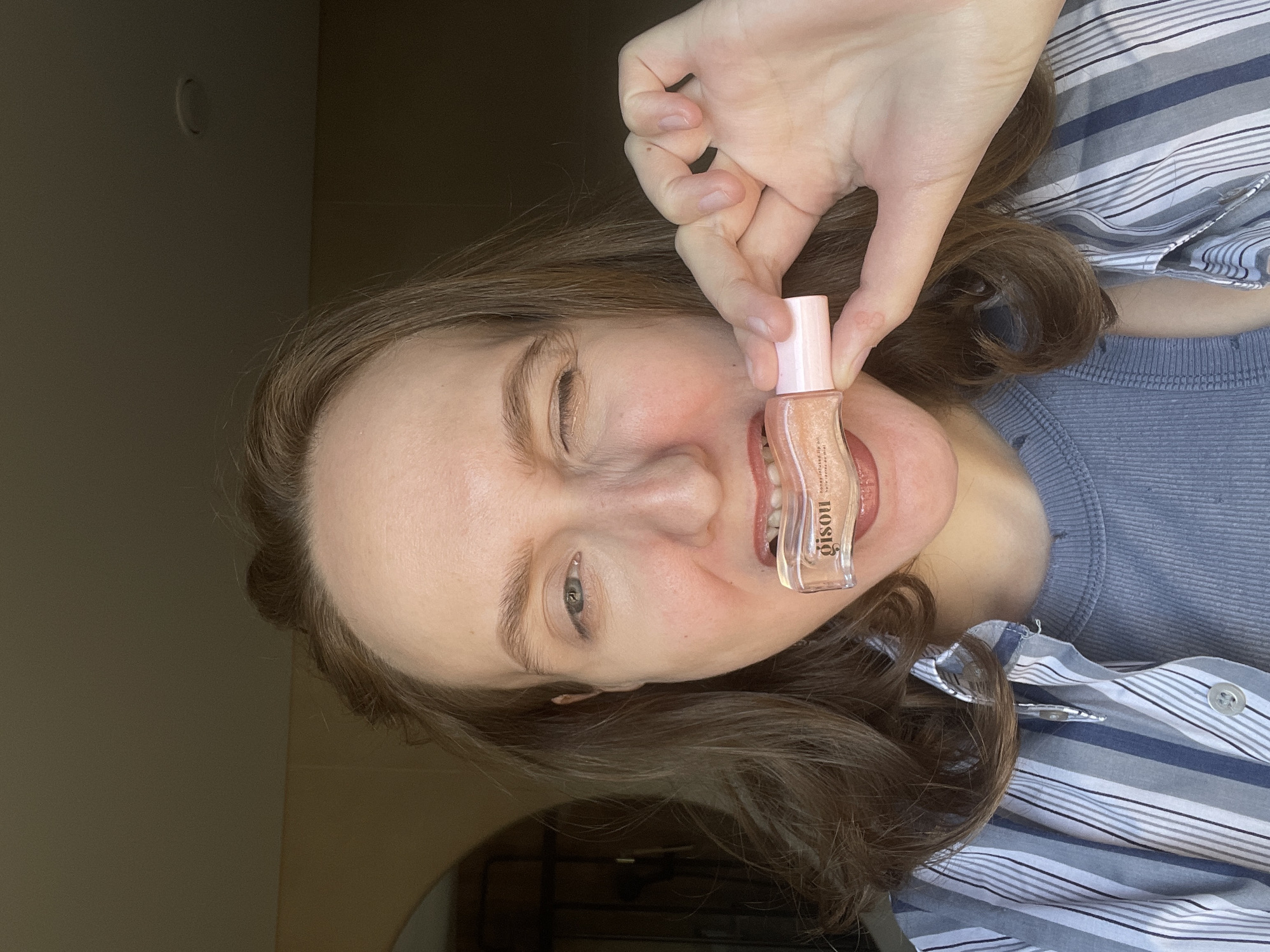 The image shows a woman holding a lip gloss tube in front of her mouth. The lip gloss tube is transparent and has pink accents on the cap and label. The woman is wearing a blue, striped shirt and a light blue undershirt. Her hair is brown and curly. The background appears to be an interior room, with a light wall visible in the lower left corner and an electrical outlet plate in the upper left corner. The image provides no evidence of being in or related to the city of Подшивалово, Russia.