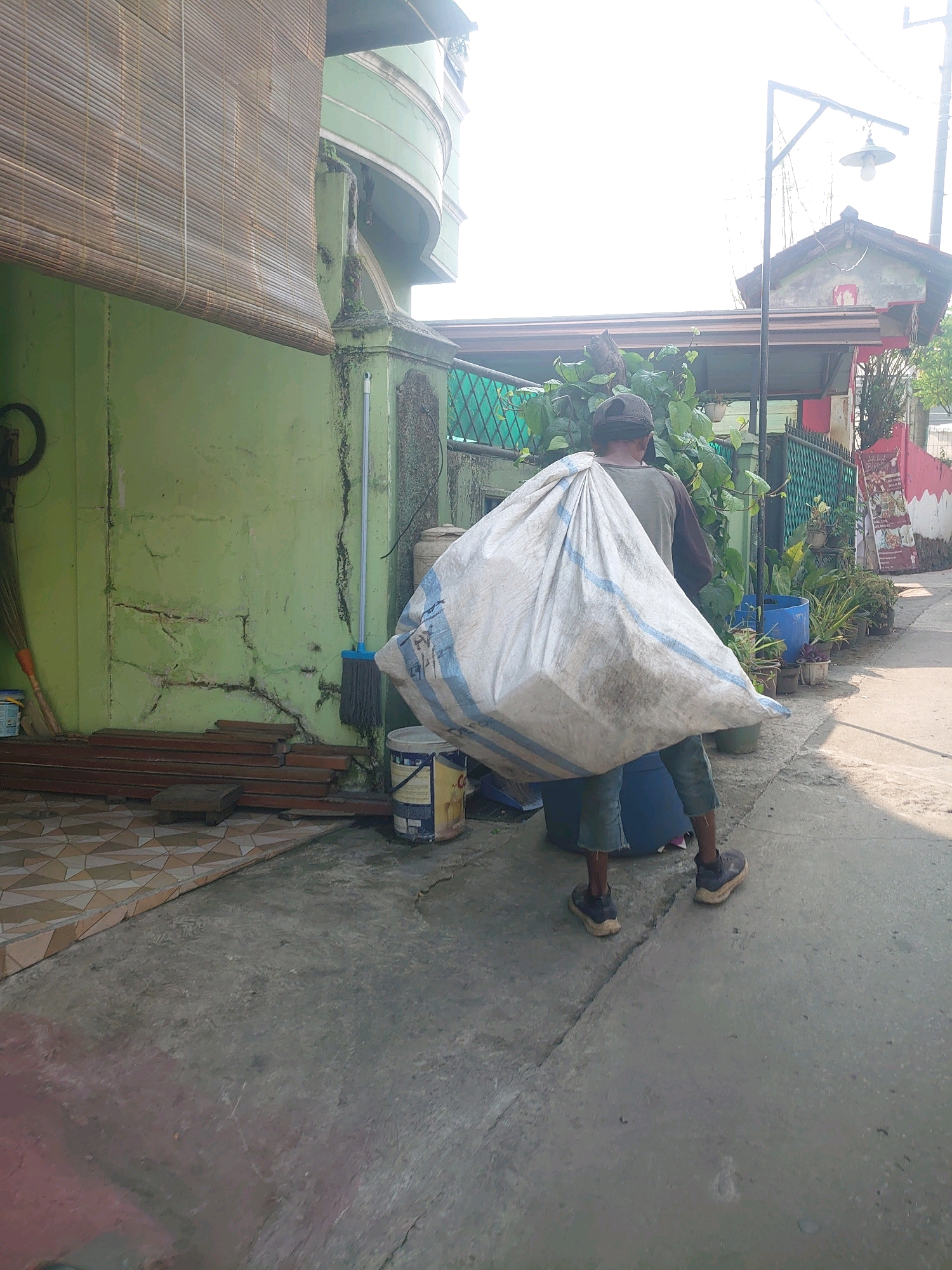 An individual is walking away from the camera, carrying a large, bulging white sack over their right shoulder. The sack has blue stripes. The person is wearing a dark long-sleeved shirt, dark shorts, and dark shoes. They are walking on a concrete pathway. To the left of the pathway is a light green wall with a bamboo shade hanging above it. There are also some metal bars stacked against the wall, and a broom leaning nearby. To the right of the pathway, there are several potted plants and a blue barrel. In the background, there is a house with a red and white painted facade and a street lamp. The scene appears to be in an outdoor, urban environment, possibly a residential area in Bojong Gede, Indonesia. The sky is bright, suggesting daylight.