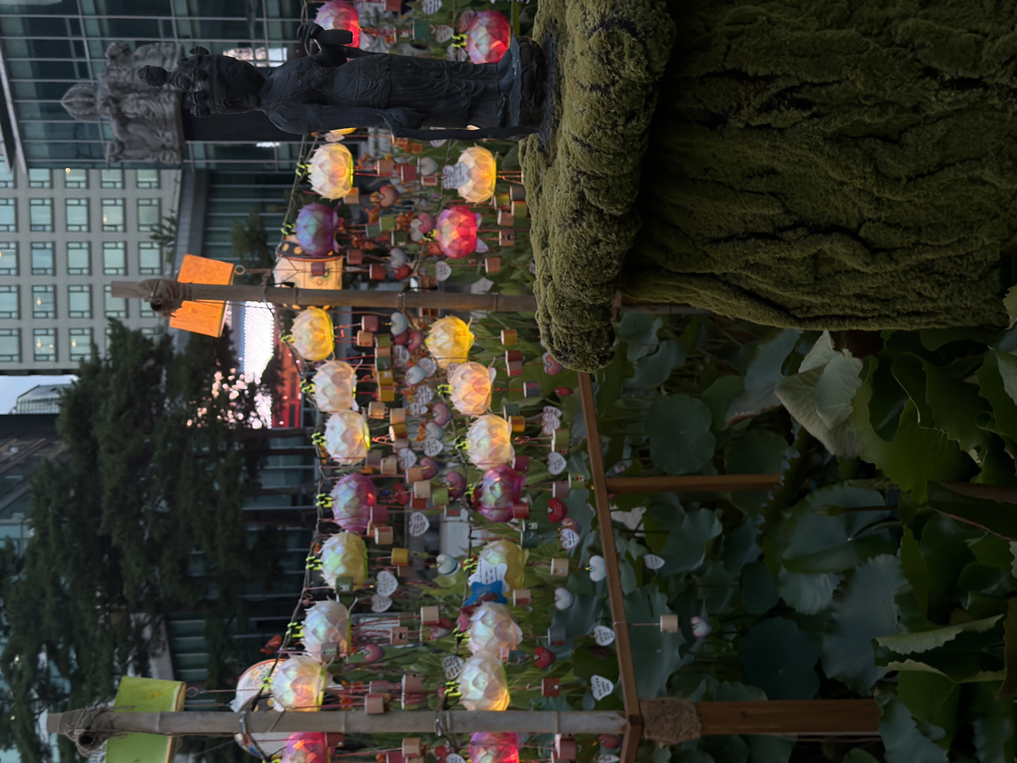 The image shows a display of lotus lanterns and prayer tags set up in a garden, likely at a Buddhist temple. In the background, modern high-rise buildings with many windows are visible, suggesting an urban setting. A dark statue of a figure wearing a crown stands prominently in the foreground, on a moss-covered pedestal. The lanterns are colorful and illuminated, casting a warm glow. Many small prayer tags, some heart-shaped, are hanging from the lanterns and a wooden structure. The overall scene appears to be a peaceful and spiritual display, possibly during a festival or special event. The presence of lotus flowers and Buddhist imagery strongly suggests this is a Buddhist temple in Seoul, South Korea.