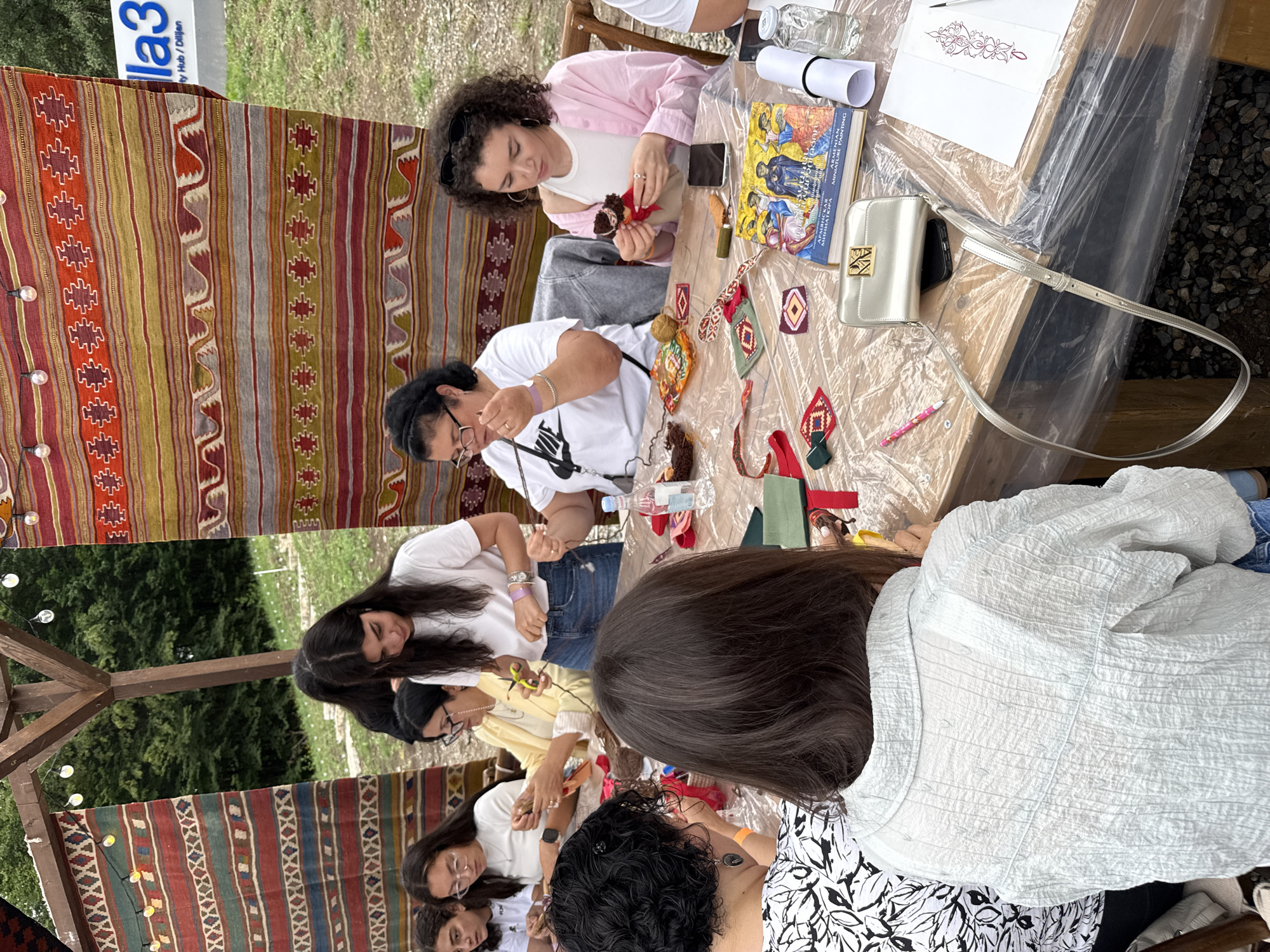 The image depicts an outdoor scene in Dilijan, Armenia, likely at a cultural event or workshop. Several people, predominantly women, are gathered around a long wooden table covered with a clear plastic sheet. They appear to be engaged in a craft activity, possibly embroidery or textile art, as evidenced by the presence of fabric, thread, needles, and small decorative pieces.
In the background, colorful woven textiles are displayed, adding to the cultural ambiance. These textiles, featuring geometric patterns and vibrant colors, hang from a wooden structure that suggests a shaded outdoor area, possibly a pavilion or an open-air market stall. The overall environment suggests a relaxed, communal setting within Dilijan, a city known for its natural beauty and artistic heritage. The people are focused on their tasks, with some holding tools and materials, while others are observing or assisting. The presence of a book titled "ARMENIAN MINIATURE PAINTING" further suggests a focus on Armenian arts and crafts.