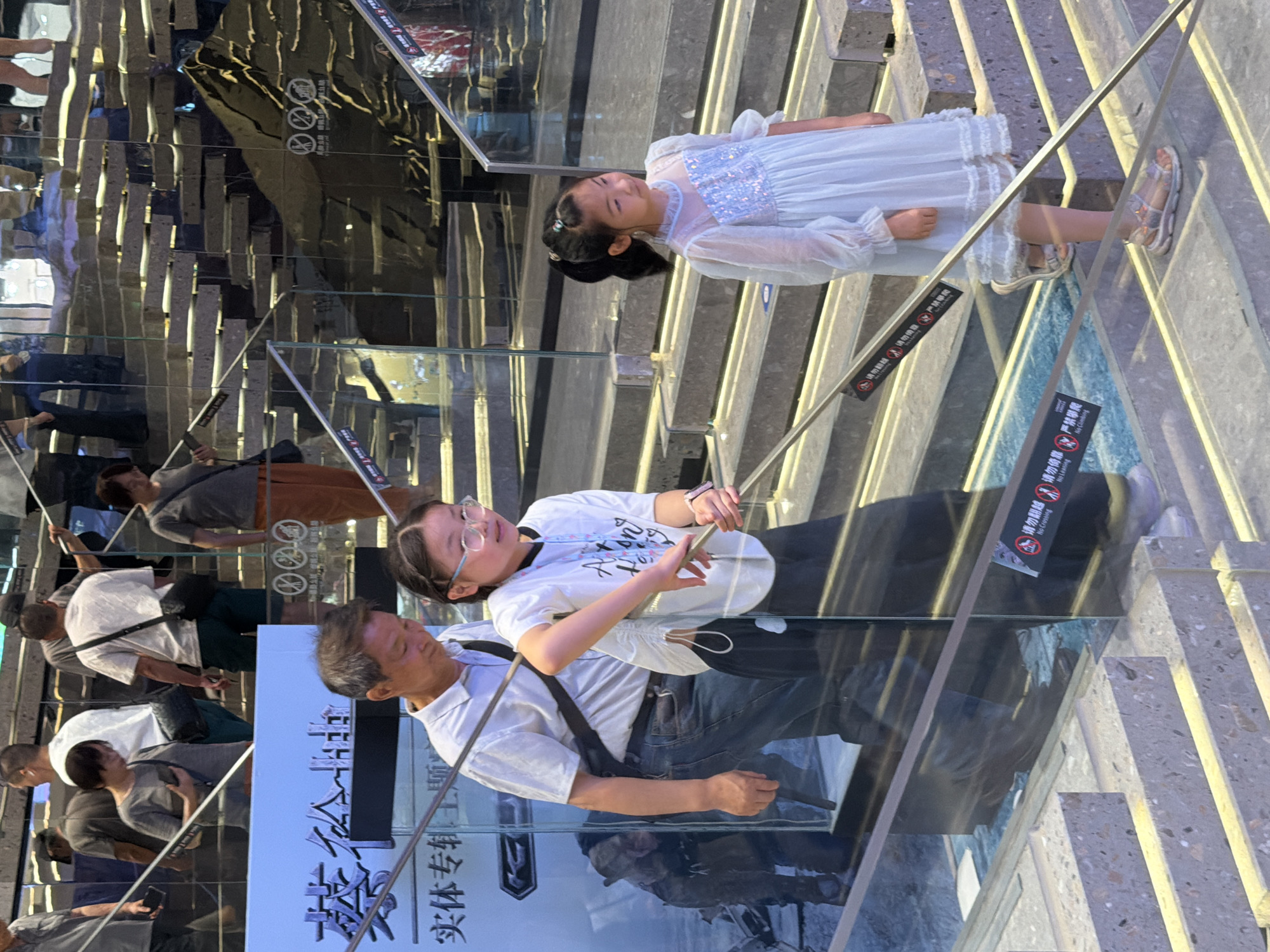In Yuzhong District, China, a father and his two daughters are ascending a modern, multi-level staircase with mirrored walls and illuminated steps. The father, wearing a white short-sleeved shirt and blue jeans, walks slightly ahead and to the left. His older daughter, in a white t-shirt and dark pants, is to his right, holding onto a silver railing. She wears glasses and has her hair pulled back. The younger daughter, in a pale blue sequined dress, walks further up the stairs to the right. She has her hair in a ponytail and wears sandals. The environment appears to be a commercial or public space, possibly a shopping mall or exhibition hall, given the presence of signage with Chinese characters and reflective surfaces. The overall scene captures a family navigating a dynamic architectural space.