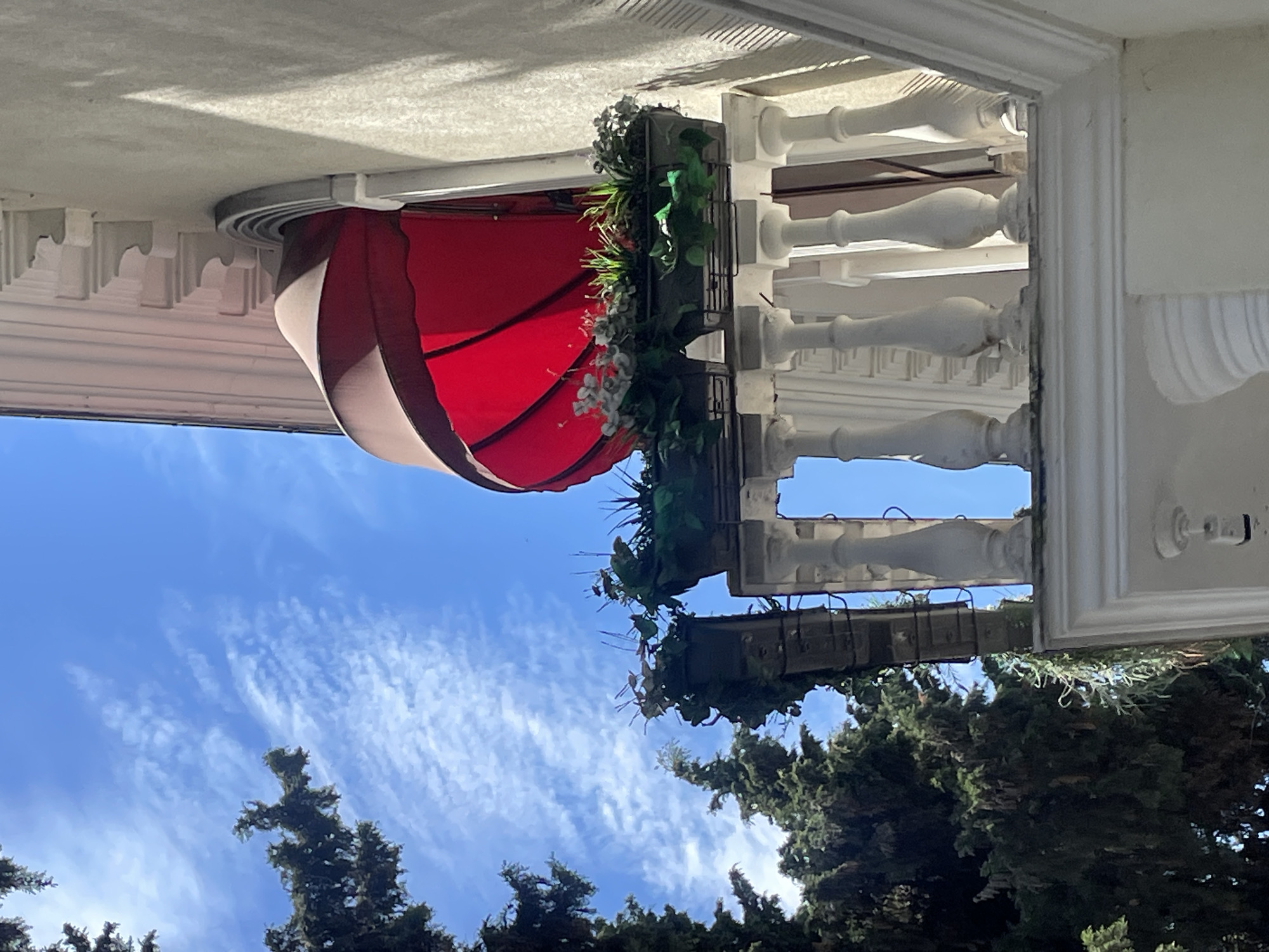 The image shows a red awning attached to a building in Sochi, Russia. The building has white columns and trim. A flower box hangs on the building with greenery. The sky is visible with some clouds and trees. The image appears to have been rotated and needs to be turned right side up.
