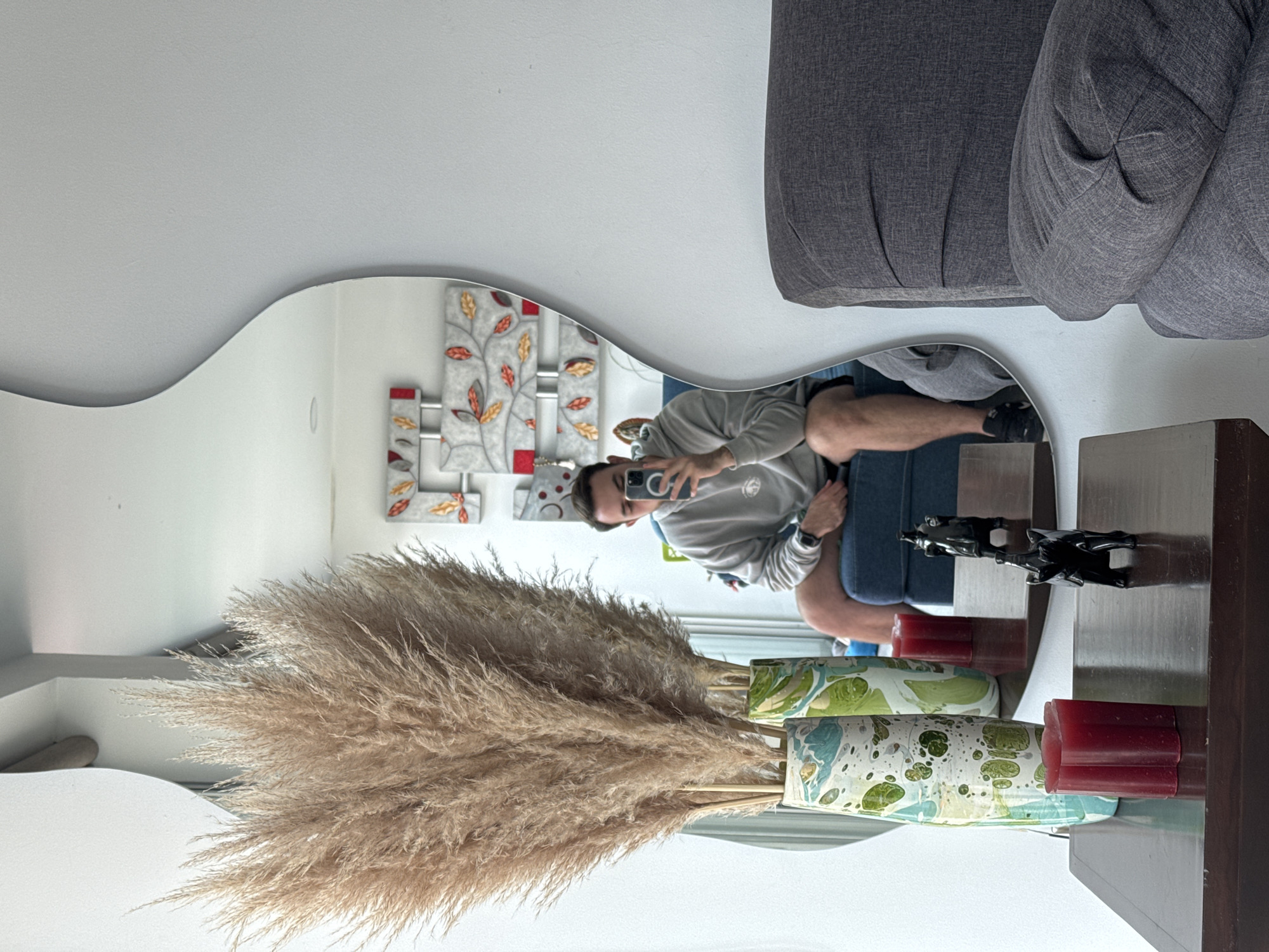 The image displays an indoor scene in San Bernardino Tlaxcalancingo, Mexico, dominated by a person taking a mirror selfie. The central subject is a male individual, reflected in a large, wavy-edged mirror, who is holding a smartphone with a noticeable 'C' or circular logo on its case to capture the photo. He is dressed in a light-colored long-sleeved shirt or hoodie, blue jeans, and a watch on his left wrist.
In front of the mirror, a substantial bundle of dried pampas grass is arranged in two decorative vases, which feature a distinctive green, blue, and white marbled pattern. To the right of these vases, two red pillar candles are visible, alongside two black horse figurines, all resting on a dark, possibly wooden, surface. A section of a grey textured sofa or armchair is positioned in the foreground, suggesting a comfortable living area. The mirror also reflects wall art behind the person, depicting a stylized tree or branch design with red, orange, and gold leaves against a grey and white background. The lighting indicates a daytime setting.