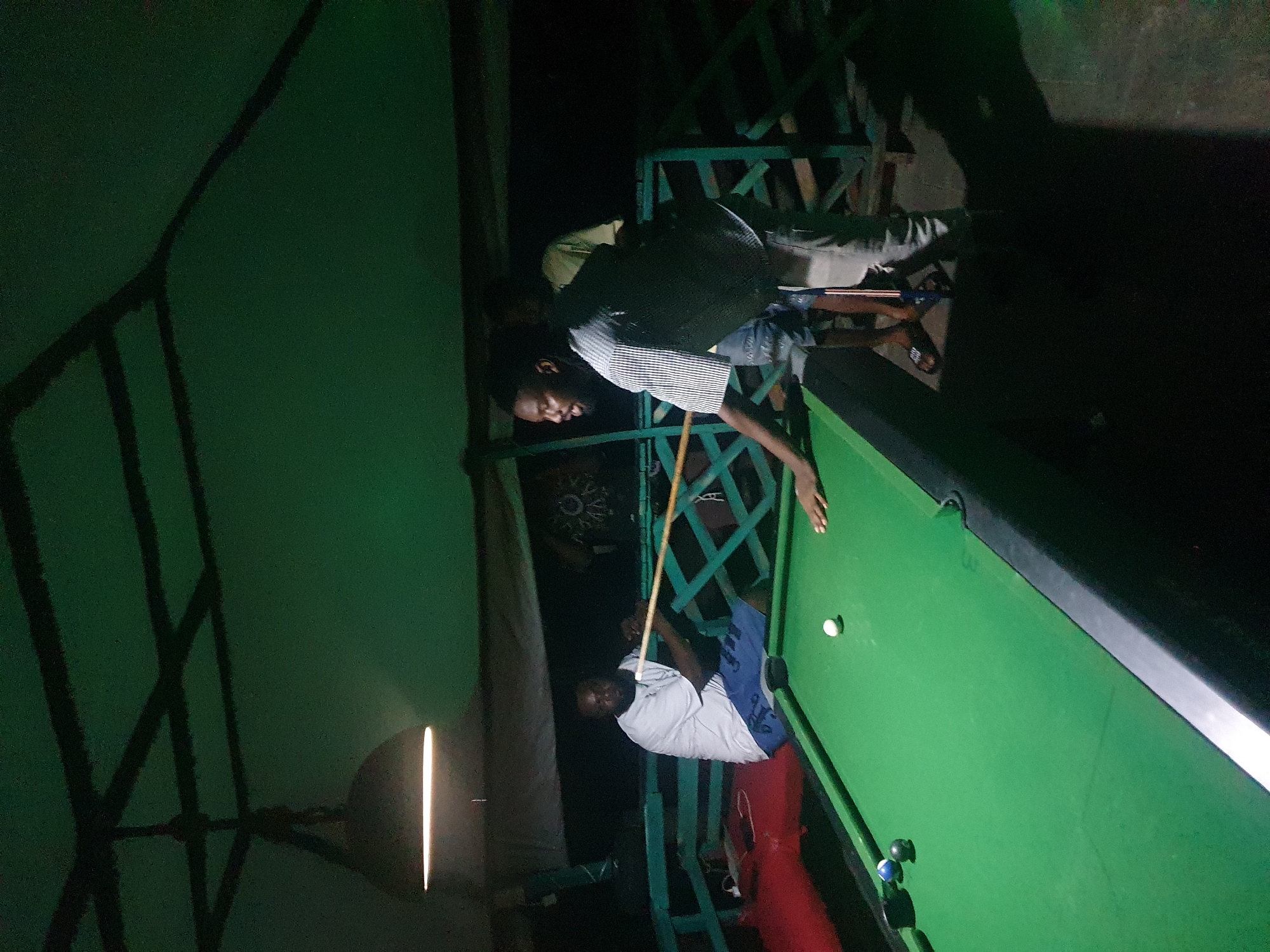 The image depicts an indoor pool hall in Lokoja, Nigeria, at night. A green pool table occupies the foreground, with several billiard balls visible, including a white cue ball. Two men are actively engaged in a game of pool. One man, seated, is wearing a light-colored t-shirt and blue shorts. The other man, standing and facing the table, is wearing a striped shirt and light-colored pants, and is in the process of striking the cue ball with a cue stick. Another person is partially visible in the background, facing away from the camera. The environment appears to be a covered area with a dark ceiling and a hanging light fixture illuminating the scene. The background suggests a casual, possibly outdoor or semi-outdoor setting with a railing and a dark, unlit area beyond.