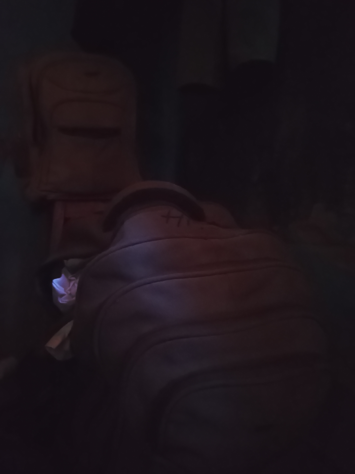 The image depicts a dimly lit scene with two backpacks prominently featured. The backpack in the foreground is a muted brown, with stitching visible across its surface. A light, possibly from a phone screen, casts a purplish glow on some crumpled paper or fabric tucked into the front pocket of this backpack. Behind it, another, similar brown backpack is positioned vertically. The setting appears to be indoors, with the low lighting suggesting it might be nighttime or a poorly lit room. The overall atmosphere is subdued and somewhat mysterious due to the limited visibility. There is no visible text in the image. The context provided places the scene in Wauro Jabbe, Nigeria, but there are no specific visual cues within the image to confirm this location. The focus is solely on the backpacks and the subtle illumination.