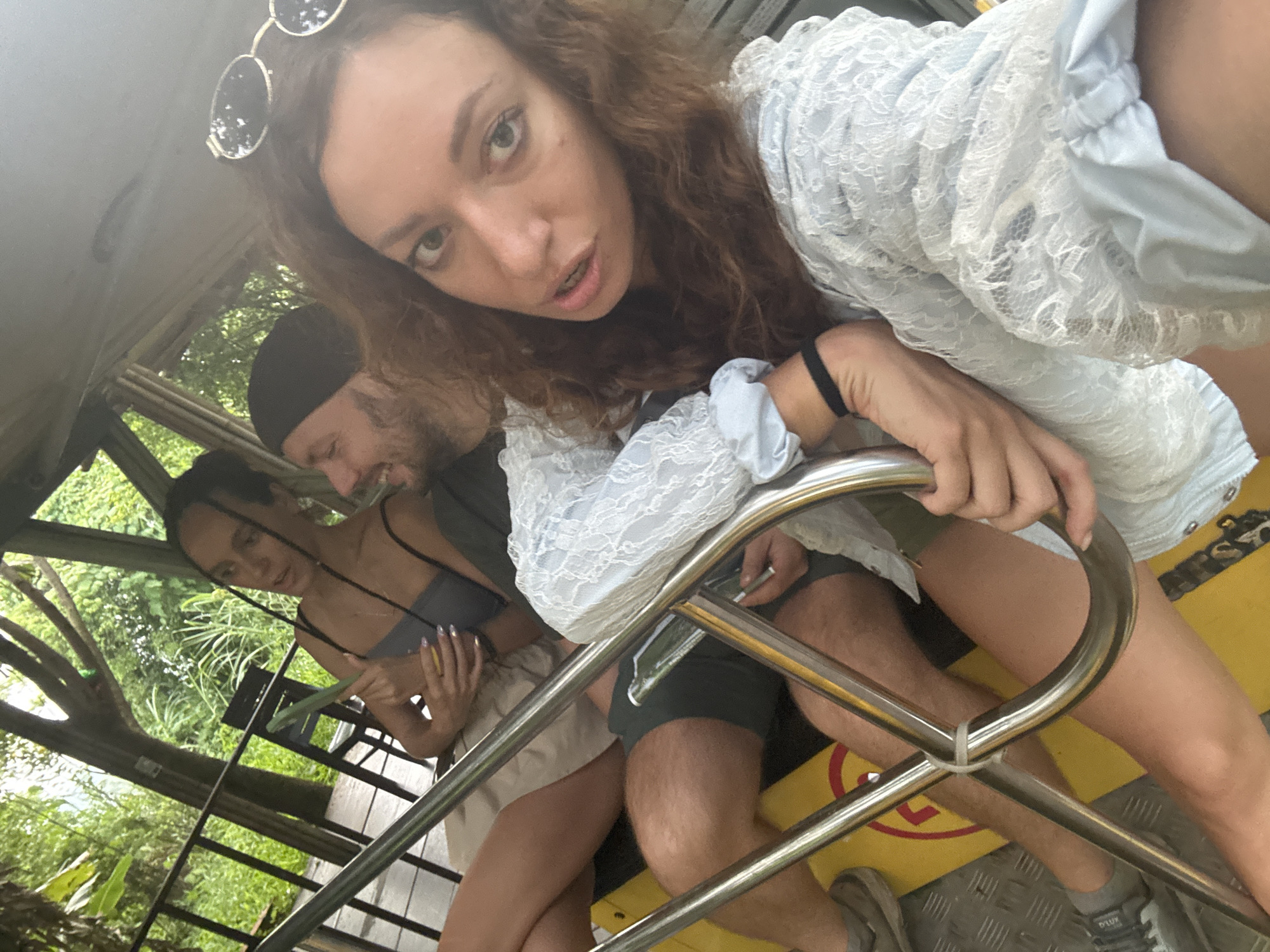 The image depicts a selfie taken from a low angle inside what appears to be a transportation vehicle. In the foreground, a young woman with long, curly brown hair is leaning into the camera, her face filling a significant portion of the frame. She is wearing a light blue, lace-trimmed top and sunglasses are perched on her head. To her left and slightly behind her, a man with a black beanie and beard is visible, looking downwards and smiling. Further in the background, another woman in a grey top and a light-colored skirt is seated, holding a yellow object. The vehicle has metal railings and a yellow surface. Lush green foliage and trees are visible through the open sides of the vehicle, suggesting an outdoor, possibly tropical or jungle-like environment. The overall impression is of a casual outing, possibly a tourist activity. The image provides no specific landmarks or architectural details that would pinpoint the exact location within Tây Hoa Lư Ward, Vietnam, beyond the general natural environment.