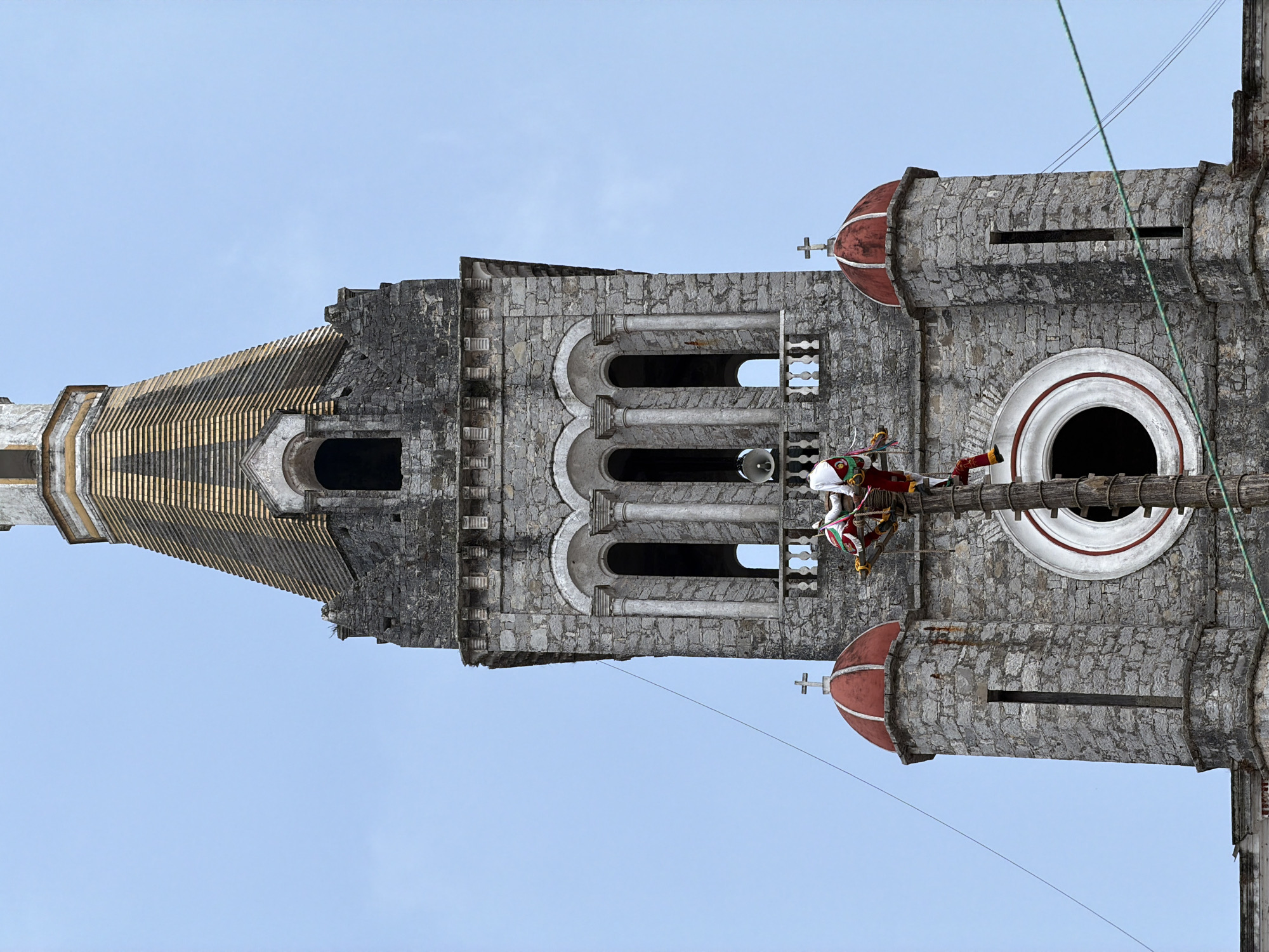 The image shows the upper portion of a tall, stone church tower in Cuetzalan, Mexico, under a clear sky. The architecture features a mix of stone types and includes arched windows, decorative columns, a dome with a cross, and a conical spire. A group of Voladores performers in traditional red and white costumes are situated on a wooden platform near the top of the tower, secured by ropes for their ritual performance.