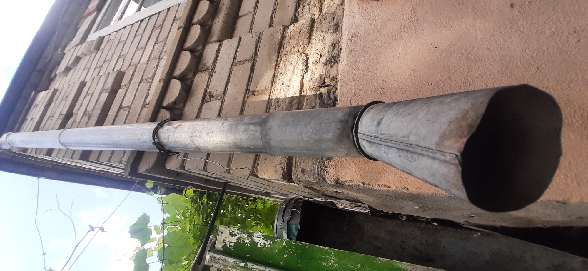 The image is a close-up, low-angle shot of a section of a brick building's exterior, focusing on a downspout. The building appears to be located in Yessentukskaya, Russia. The downspout is made of galvanized metal and runs vertically down the side of the building. It is secured to the brick wall with wire. The brickwork is light tan and shows signs of age and some damage, with a decorative band of semicircular brickwork near the top. To the left of the downspout, there is a glimpse of green foliage, possibly from a vine, and a portion of a weathered green structure. The sky is visible in the upper left corner, with some clouds. The lower part of the downspout widens into a flared opening. The overall scene suggests an outdoor, possibly residential or older commercial, setting. No people are visible in the image.
