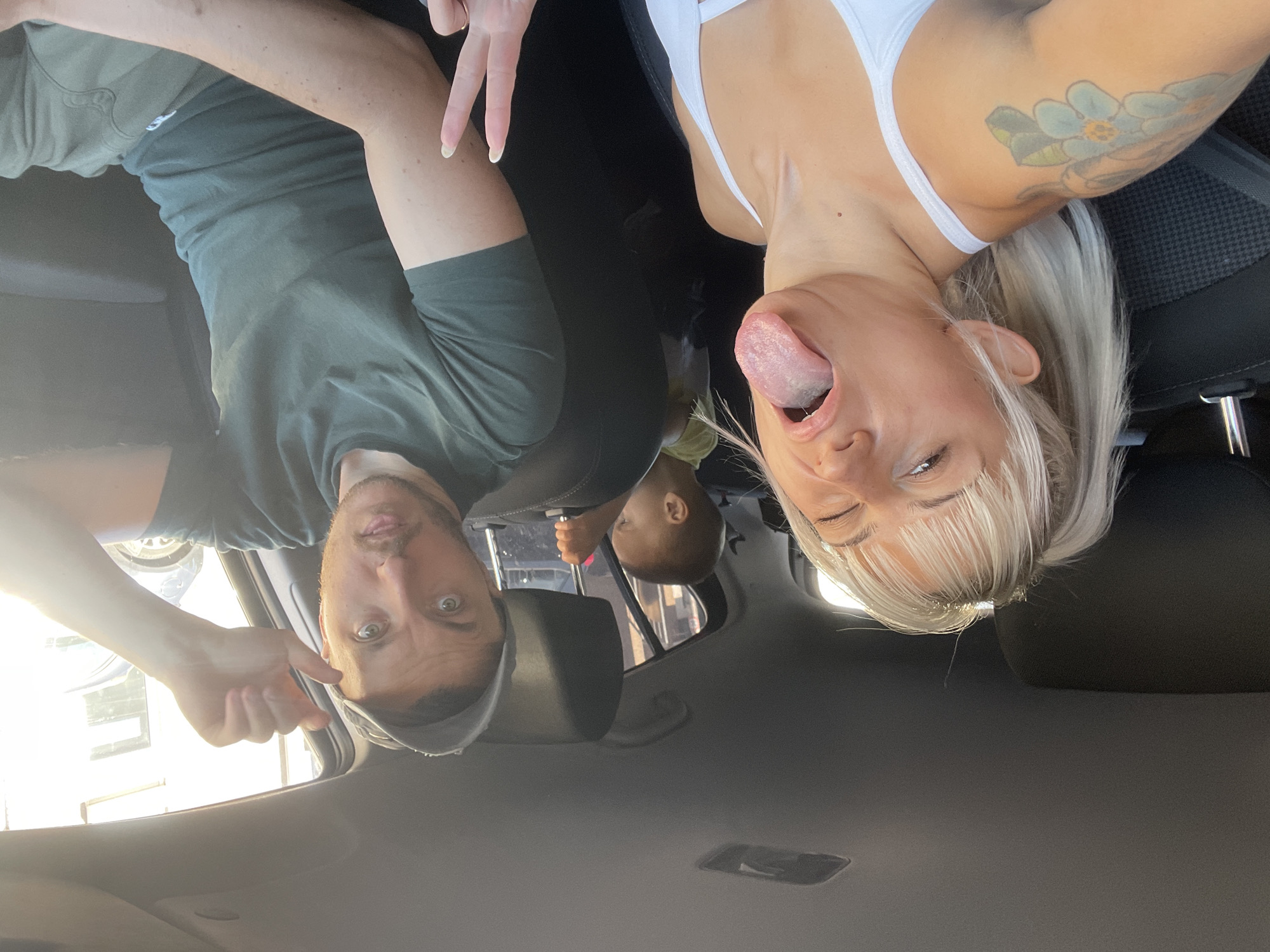 The image depicts three individuals inside a car. The photograph appears to be taken from the car's ceiling looking downward. On the left, a man is visible wearing a green shirt, and on the right, a woman wearing a white top is sticking her tongue out. A child is in the back seat. The overall setting is a car interior. Based on the exterior view through the car windows, the location is in a city, possibly Tilburg, Netherlands, as it was mentioned in the prompt. However, there isn't enough evidence in the image to confirm the exact location.