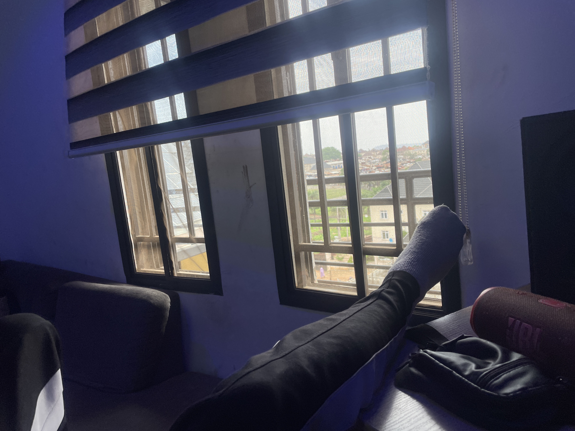The image captures an indoor scene, likely a living room or bedroom, illuminated by natural light entering through two windows on the right and artificial blue light on the left wall. A person is relaxing, visible by their legs stretched out towards the windows. They are wearing black pants and a white or light-colored sock on their visible foot.
The windows feature horizontal striped blinds, alternating between dark blue and lighter, translucent fabric. Outside the windows, security bars are visible, and beyond them, an urban or suburban landscape unfolds with various buildings and some greenery under bright daylight.
Inside the room, to the left, a dark-colored sofa or couch is partially visible. On the right, next to the person's extended leg, sits a red or maroon JBL portable speaker with "JBL" clearly visible on its side. A black zippered pouch or bag is placed near the speaker on a dark surface. The wall on the right has a dark, possibly electronic, object or screen partially visible. A pull cord for the blinds hangs vertically between the windows. The overall impression is one of relaxation during the daytime in a domestic setting in Abuja, Nigeria.