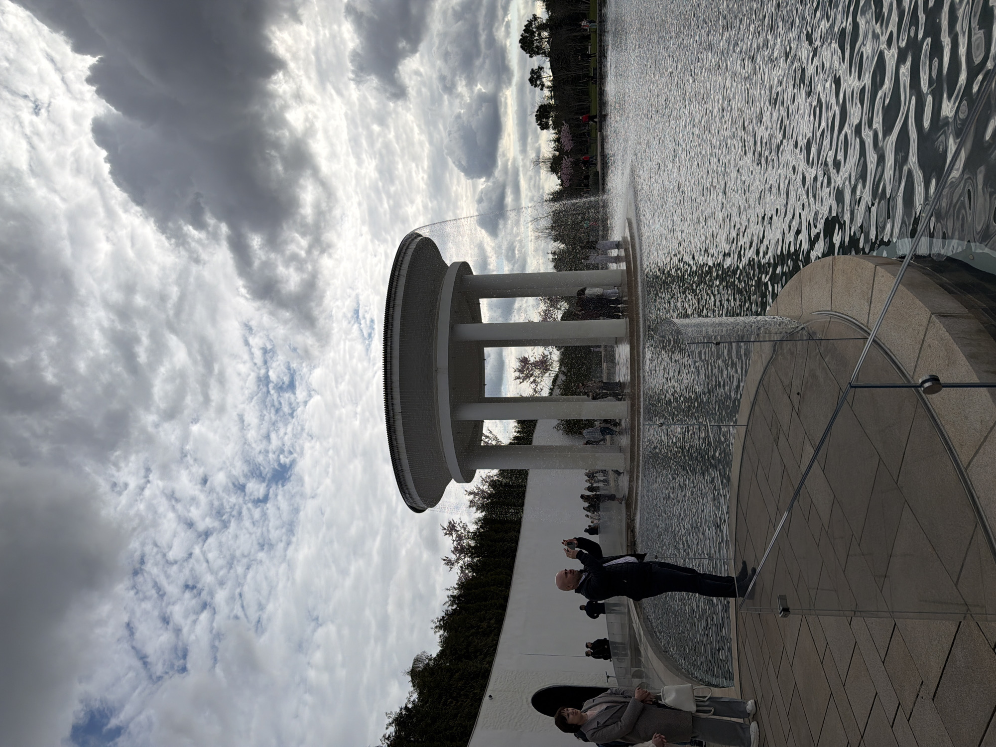The image shows a circular, colonnaded structure in a body of water. The sky is overcast with varying cloud formations. People are visible around and within the structure, some are standing, and some are taking photographs. One man is holding a camera. The ground surrounding the water is paved. The location is likely a memorial or fountain in a public space in the city of Krasnodar, Russia. Vegetation, including trees and bushes, lines the background.