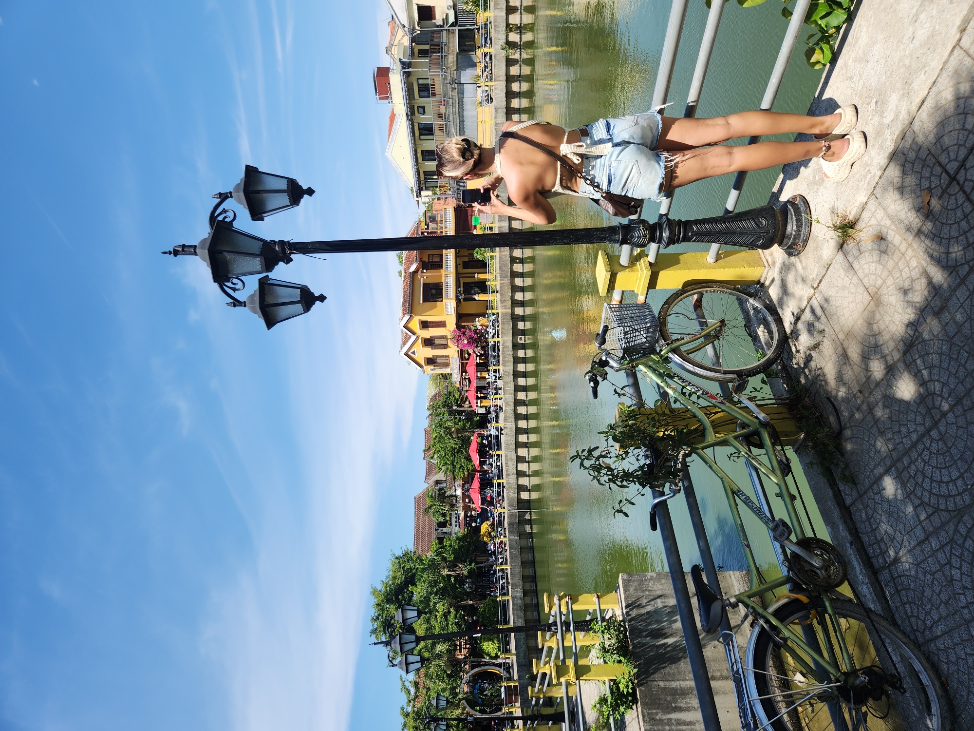 A young woman with blonde hair pulled into a bun stands on a paved walkway, facing away from the camera, and takes a photo with her phone of the scenery across a river in Hoi An, Vietnam. She is wearing a white crocheted halter top and light blue denim shorts. Her left shoulder is adorned with a brown purse strap. She wears light beige slip-on sandals. Beside her, to the left, is a black lamppost with three lights. In the foreground, to the left of the lamppost, is a light green bicycle with a wire basket on the handlebars. The bicycle is leaning against a yellow railing that borders the walkway. The walkway itself is paved with interlocking circular stones, casting dappled shadows from the lamppost and foliage. Across the river, the opposite bank features a row of two- and three-story buildings with yellow facades and red tiled roofs. Red umbrellas are open outside some of the buildings, suggesting outdoor seating for businesses. Lush green trees are visible behind the buildings. The river is a calm, greenish-brown color and reflects the blue sky and surrounding structures. The sky is a clear, bright blue with a few wispy white clouds. The image captures a sunny day in a picturesque, canal-side area of Hoi An.