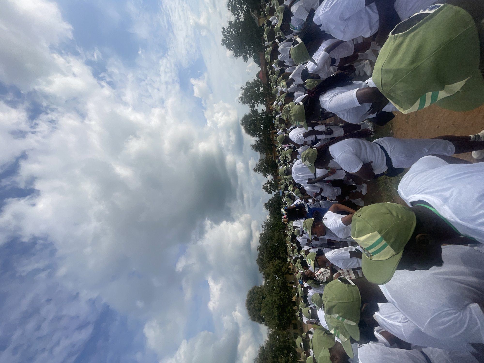 The image depicts a large gathering of young adults, predominantly wearing white t-shirts and green caps with white and green stripes. They are assembled outdoors on a dirt ground, with a line of trees and some low-rise buildings visible in the background. The sky above is partially cloudy. The scene suggests an organized event or activity involving a significant number of participants, likely in a rural or semi-urban setting. The presence of matching uniforms implies a specific group or program. The location is identified as Ise- Ekiti, Nigeria.