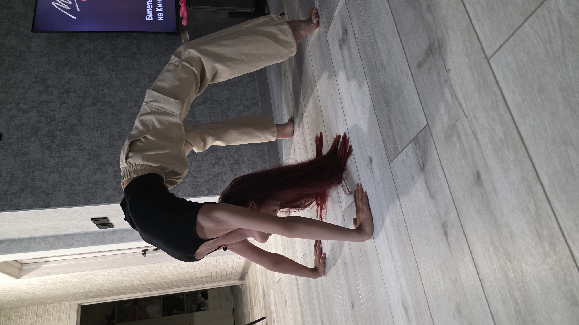 A woman with long, auburn hair is performing a backbend, with her hands and feet placed on a light-colored wooden floor. She is wearing a black tank top and loose-fitting tan pants. The background includes a grey wall with a television mounted on it, and a section of brick-patterned wall with a doorframe. The location is indoors and appears to be a domestic setting, not necessarily specifically in Bulgakovo, Russia. There is no definitive evidence within the image to verify it being located in Bulgakovo.