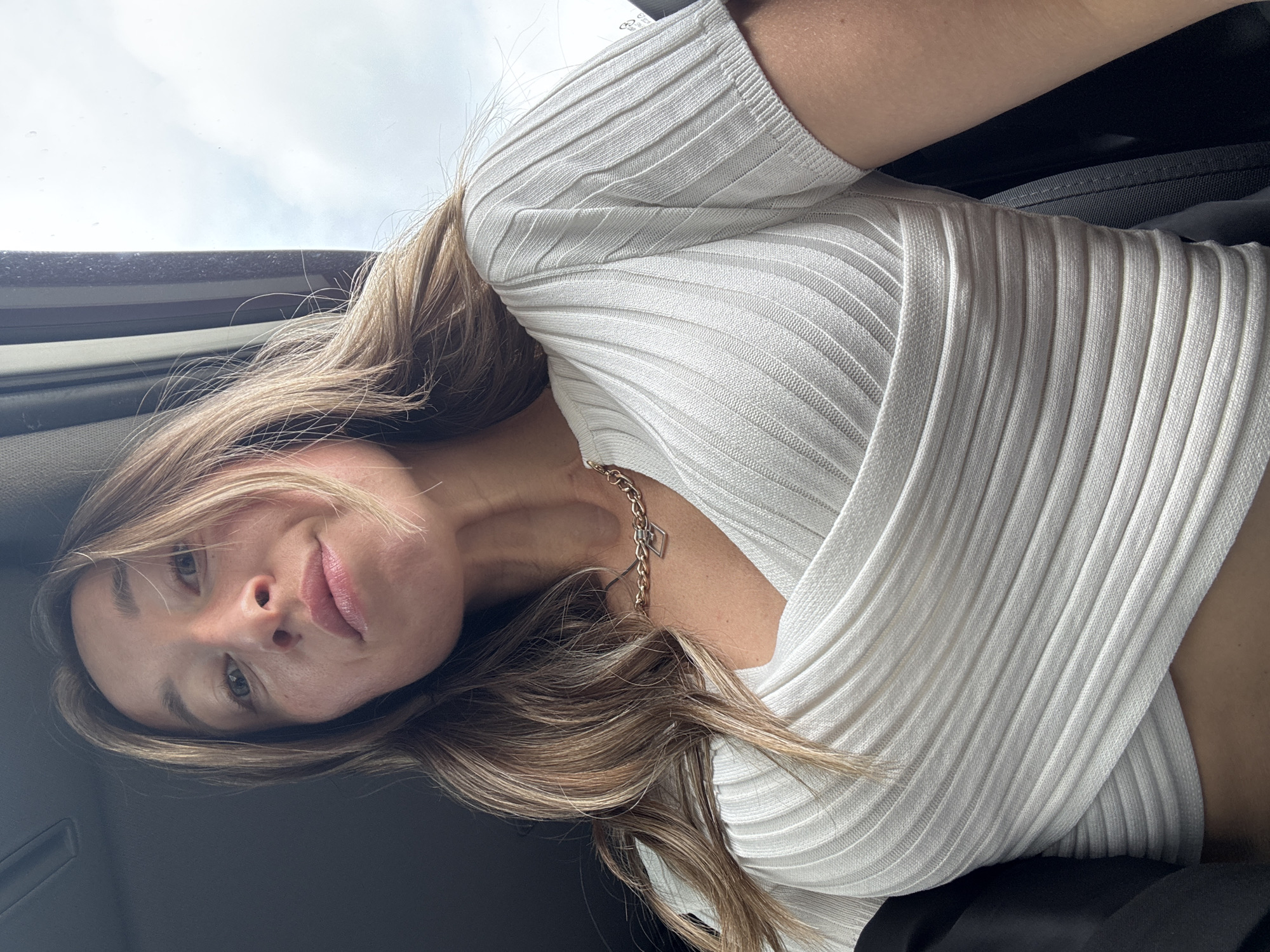 The media file displays a young woman inside what appears to be a vehicle, likely a car. She is positioned for a low-angle shot, looking directly at the camera with a soft, engaging smile. Her head is tilted to the side, and her long, light brown hair with blonde highlights frames her face and shoulders.
She is wearing a light-colored, possibly white or off-white, ribbed long-sleeve top, which features a V-neck and a textured pattern. Around her neck, a delicate gold-colored chain necklace with a small, geometric (possibly rectangular or diamond-shaped) charm is visible.
The setting suggests she is in the passenger or driver's seat of a car. Elements of the car's interior, such as dark upholstery or trim, are visible in the background and foreground. Above her, a window or sunroof reveals a bright, overcast sky, indicating it is daytime with cloudy weather. No distinct text is visible within the image. This scene is contextualized as being in Chaikovsky, Russia.