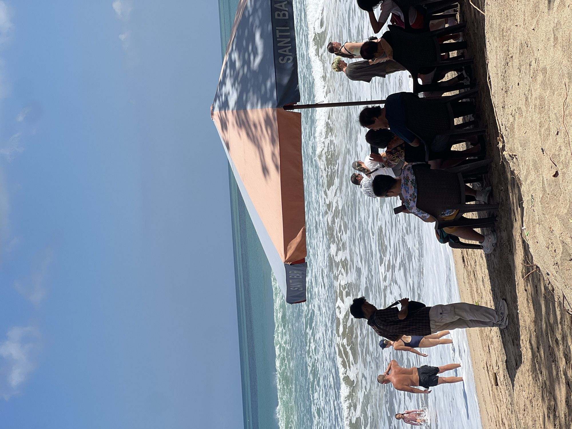 In Kuta, Indonesia, a beach scene unfolds under a bright, partly cloudy sky. A large, striped umbrella, colored coral and gray, casts dappled shadows on the sand, with "SANTI BAR" visible on its edge. In the foreground, people are seated at tables under the umbrella, facing the ocean, some appearing to be engaged in conversation. To the left, near the water's edge, a man in a plaid shirt stands, possibly interacting with someone off-camera, while a woman in a dark swimsuit and a man in black swim shorts are in the shallow water. Further out, waves crash onto the shore. The environment is a sandy beach with the turquoise ocean stretching to the horizon.