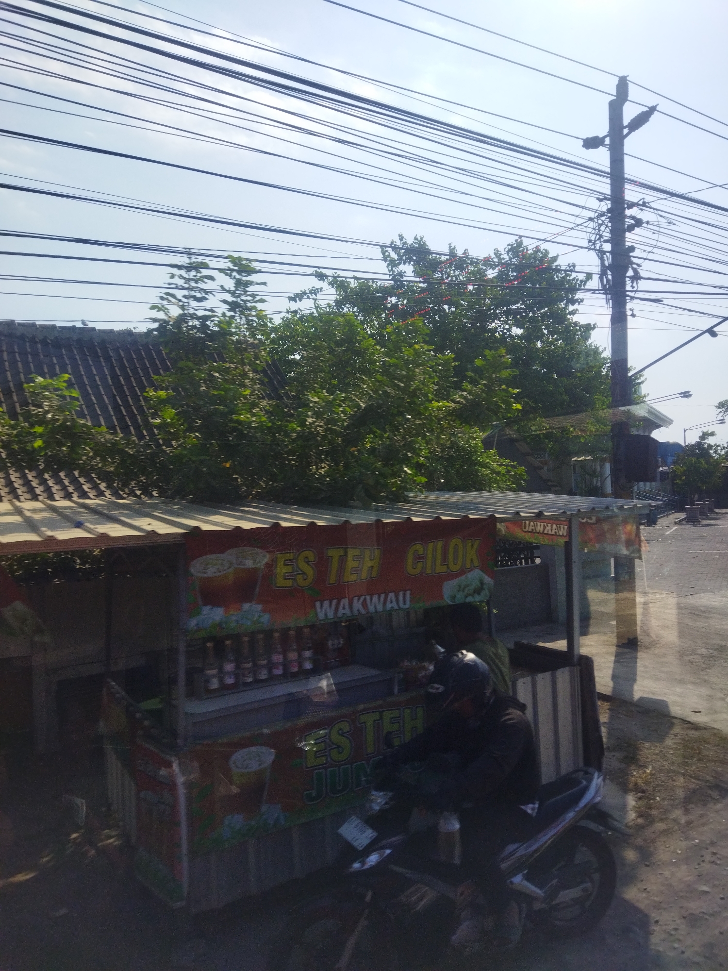 In Nguter, Indonesia, a street vendor stands behind a cart selling drinks, primarily iced tea. The cart is adorned with colorful banners displaying images of iced tea and text that reads "ES TEH CILOK" and "WAKWAU". Various bottled drinks are neatly arranged on shelves. A motorcycle is parked beside the cart, and a person wearing a helmet is seated on it, appearing to be a customer or an associate of the vendor. The scene is set against a backdrop of lush green trees, a tiled roof of a building, and a clear sky crisscrossed with numerous electrical wires. The ground is paved with asphalt, and the overall atmosphere suggests a sunny day, likely in a semi-urban or roadside setting.