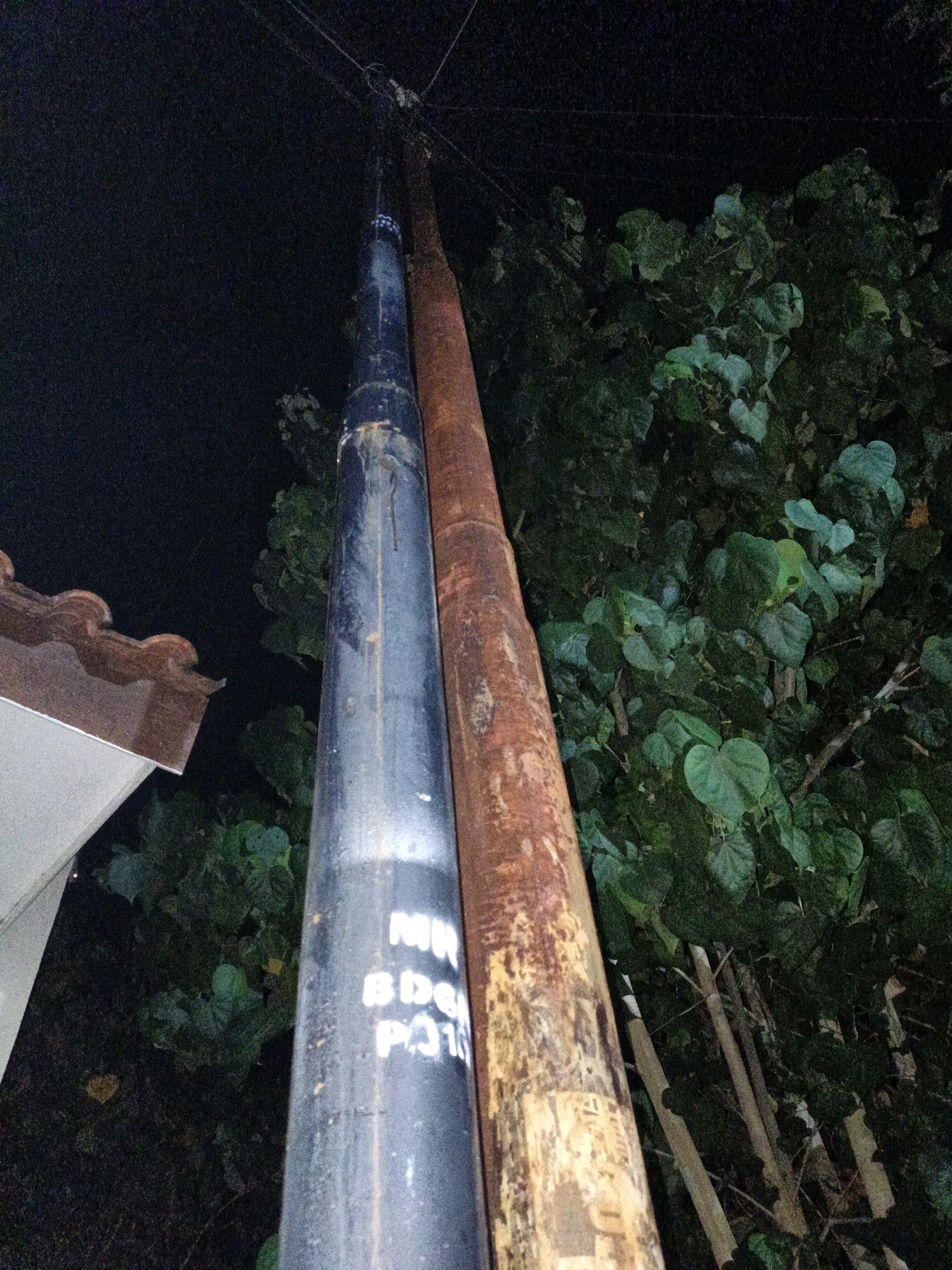 Two utility poles stand adjacent to each other at night in Cikal, Indonesia. The pole on the left is painted dark blue and shows white lettering that reads "MR BDC PG16". The pole on the right is weathered and appears to be made of a reddish-brown material, possibly rusted metal or a weathered tree trunk. Both poles extend upwards and are connected to overhead wires. To the left of the poles, a portion of a building with a tiled roof is visible. Behind the poles, dense foliage from trees is present, illuminated by an unseen light source. The ground is dark and indistinct.