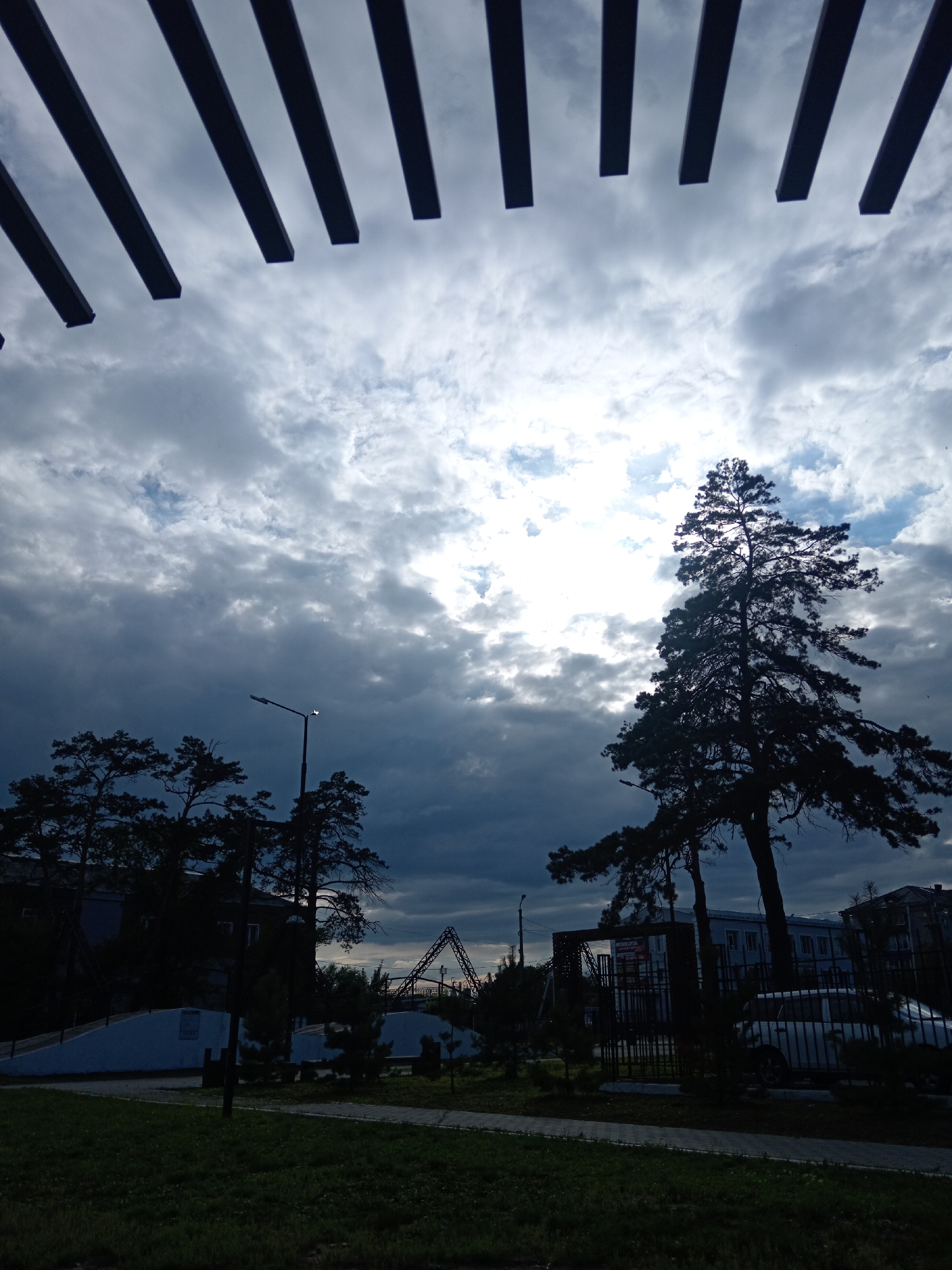 The image depicts a street scene in Belogorsk, Russia, under a cloudy sky with patches of sunlight. In the foreground, a grassy area is visible, with a paved walkway and a low concrete structure. A street lamp stands to the left of the frame, silhouetted against the sky. Several trees, also silhouetted, are scattered throughout the middle ground. Behind the trees, buildings and a playground structure are visible. A white car is parked to the right of the frame. The overall impression is one of a somewhat overcast day in a residential area.