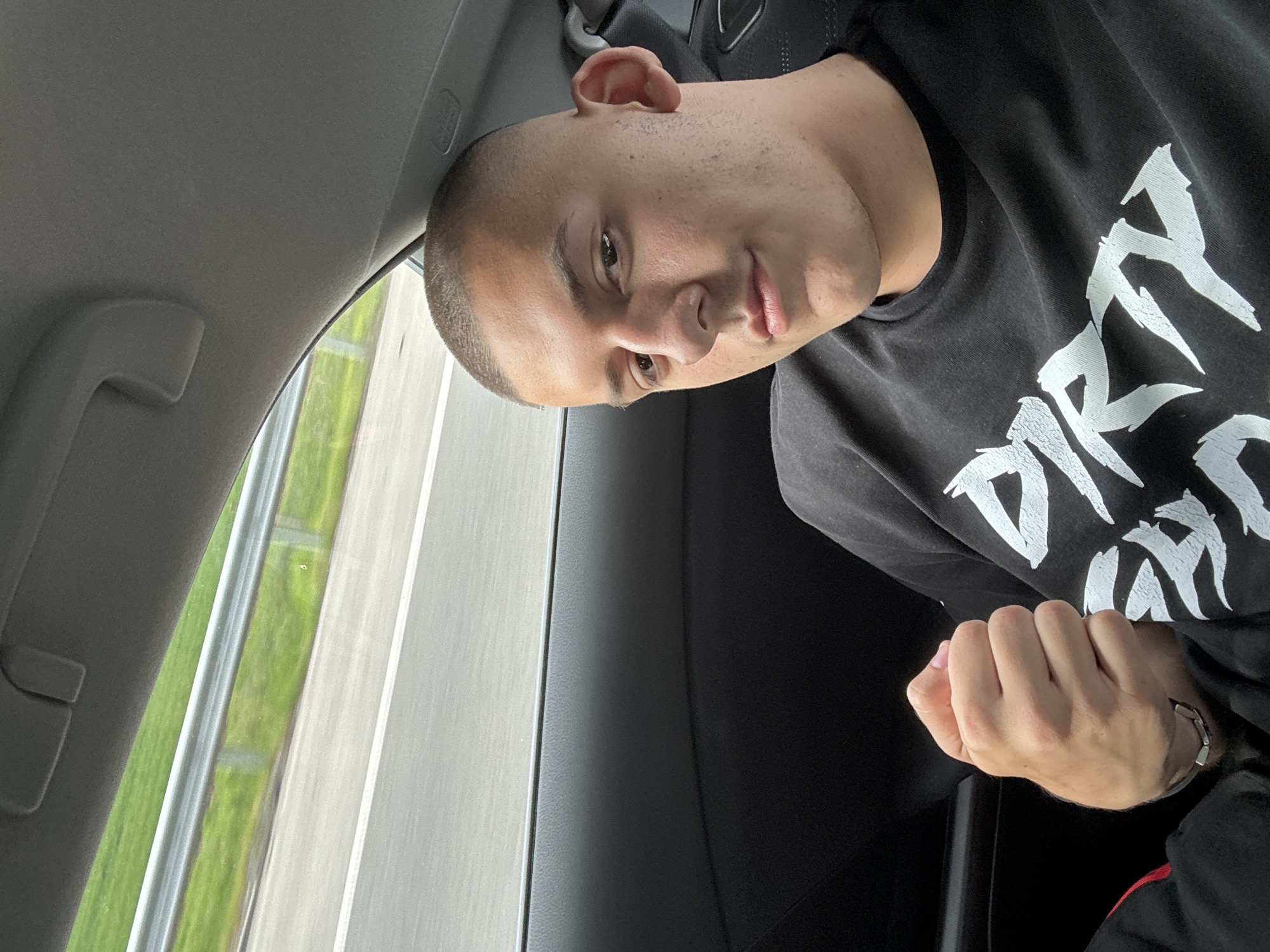 A man with short-cropped hair is in the passenger seat of a car, posing for a photo. He is wearing a dark-colored long-sleeved shirt with a white graphic print. His right hand is clenched in a fist. A car window shows passing scenery consisting of a road and greenery. The location in the city of Svoboda, Bulgaria, is not identifiable based on the visual content of the image.