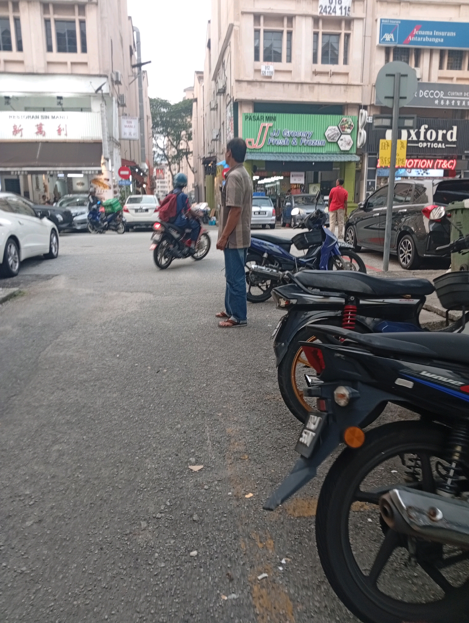 The image depicts a street scene in Selayang Municipal Council, Malaysia, characterized by a row of multi-story buildings lining the right side of the street. The architecture suggests a commercial area with various shops and businesses. On the left, a white car is parked, and other vehicles, including motorcycles and cars, are visible on the road, indicating moderate traffic.
In the foreground, several motorcycles are parked, with one prominently displayed in the lower right portion of the frame. A dark-colored motorcycle with a red shock absorber is positioned behind it. A person wearing a blue helmet and a backpack, possibly a delivery person, is riding a blue motorcycle in the middle of the street, moving away from the viewer. Another person in a red shirt and beige pants is standing near a black car, facing away from the camera. A man in a light-colored shirt and blue jeans is standing on the sidewalk, looking towards the street.
The buildings have a muted color palette, with signs in various languages, including English and Chinese, indicating commercial signage. One sign reads "RESTORAN SIN MANU" in English and Chinese characters. Another sign displays "PASAR MINI JJ Grocery Fresh & Frozen." Further down the street, a sign for "OXFORD OPTICAL" is visible. The road surface is asphalt, appearing worn in places, with some debris scattered on it. The overall lighting suggests it is daytime, possibly late afternoon given the soft light. The environment appears to be a typical urban commercial street.