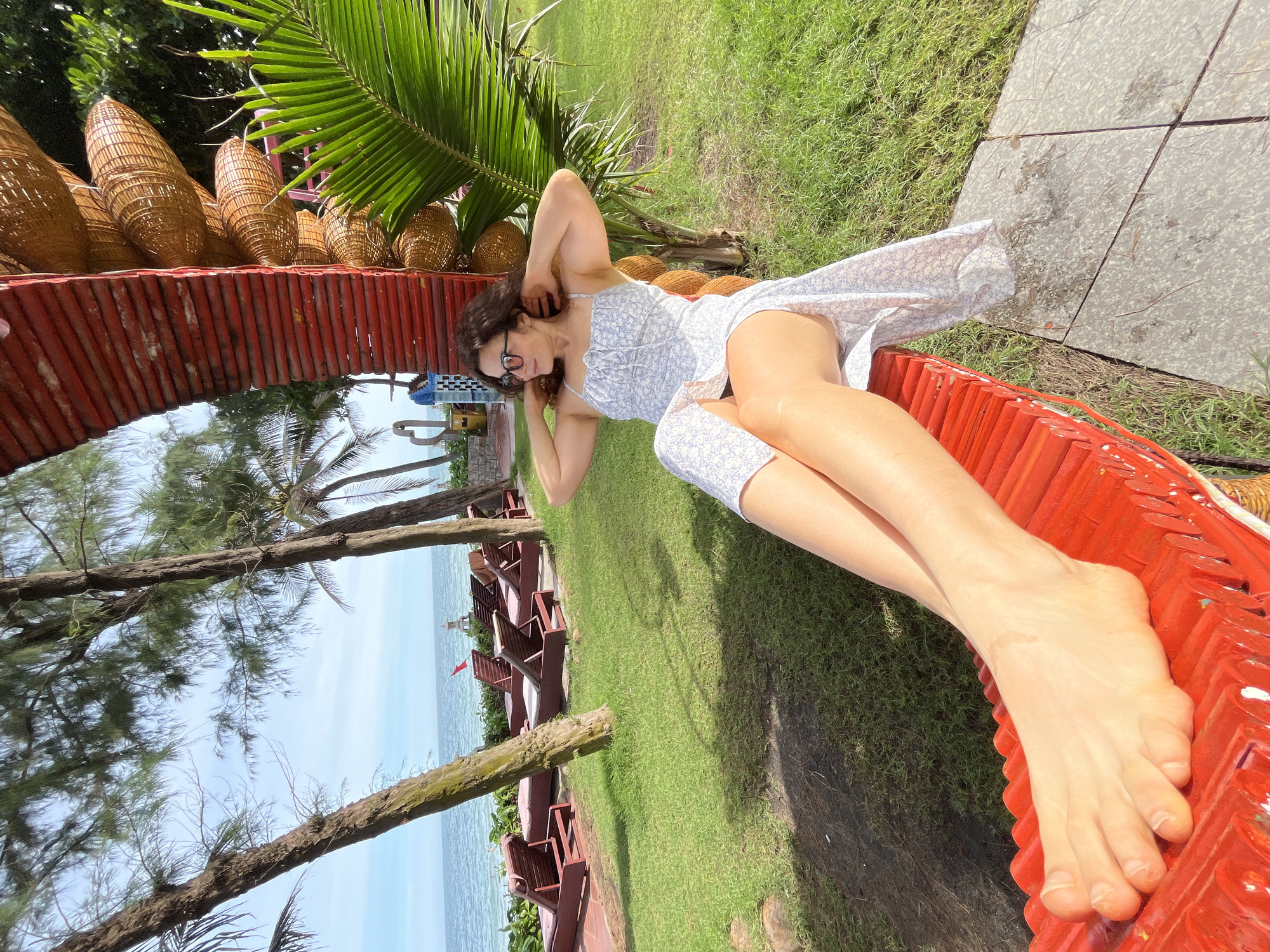 A woman is reclining on a bright red bench in a grassy area with palm trees and the ocean in the background. She is wearing a light-colored floral dress and sunglasses, with her hands behind her head. The bench appears to be part of a larger, uniquely constructed seating area that includes woven basket-like elements. In the background, lounge chairs are arranged on a paved area near the ocean, and a red flag is visible near the water. The setting is outdoors and appears to be a tropical resort or beachfront location, consistent with Mũi Né, Vietnam.