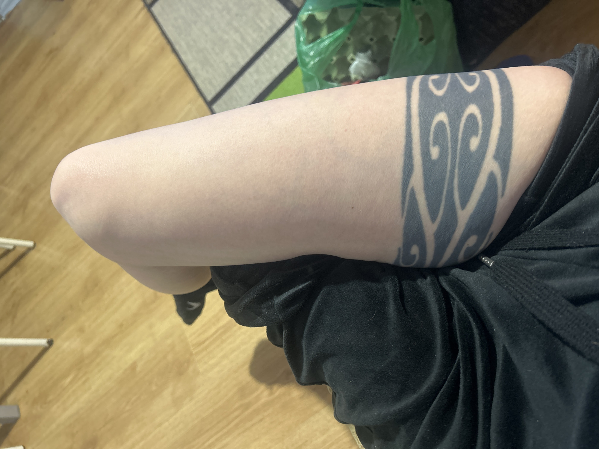 A high-angle close-up shows a person's right leg crossed over their left knee. The person is wearing black sweatpants. A tribal-style tattoo encircles the upper part of the right leg. Part of a wooden floor is visible in the foreground and background. A gray, black, and white rug is in the background, along with the corner of a green plastic bag containing a white object. A small section of a chair leg is visible in the lower left corner. The location is Unknown, Unknown.