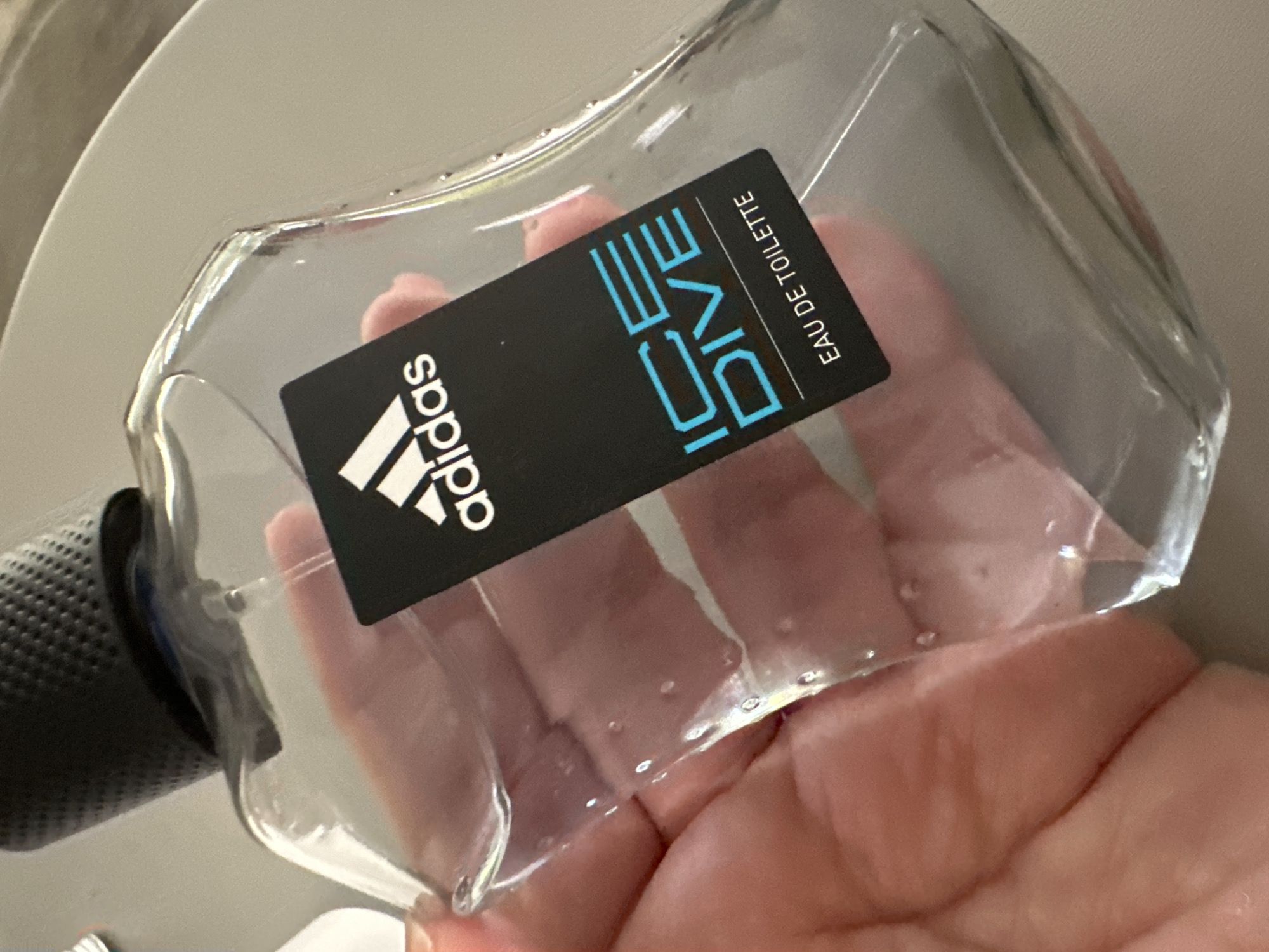 A hand is holding an Adidas "Ice Dive" Eau de Toilette perfume bottle. The bottle is clear glass, with a black cap that has a textured grip. The label on the bottle is black with white Adidas branding and blue text that says "ICE DIVE" and "EAU DE TOILETTE". The image appears to be taken indoors, possibly in a bathroom or dressing room, given the presence of a reflective surface that could be a mirror or countertop, and the lighting conditions. There are no identifiable landmarks or specific details that would indicate a location in Krefeld, Germany.