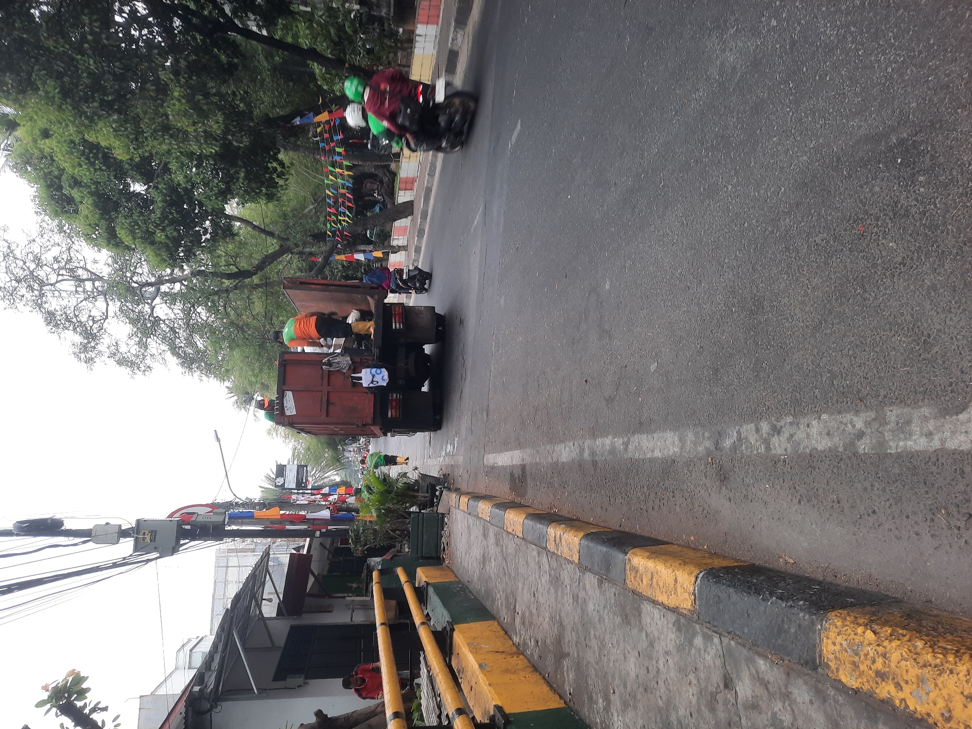 A red garbage truck with workers is parked on the side of a street in North Jakarta, Indonesia. Two workers in orange vests are visible in the back of the truck, and one is on the side, appearing to be working. A man in a red shirt is standing near a yellow and black striped barrier on the sidewalk. A few motorcycles are on the road; one carrying two passengers in helmets, and another with a single rider. The street is lined with trees and buildings on one side, and flags are strung across the street. The sidewalk has a yellow and black striped curb.