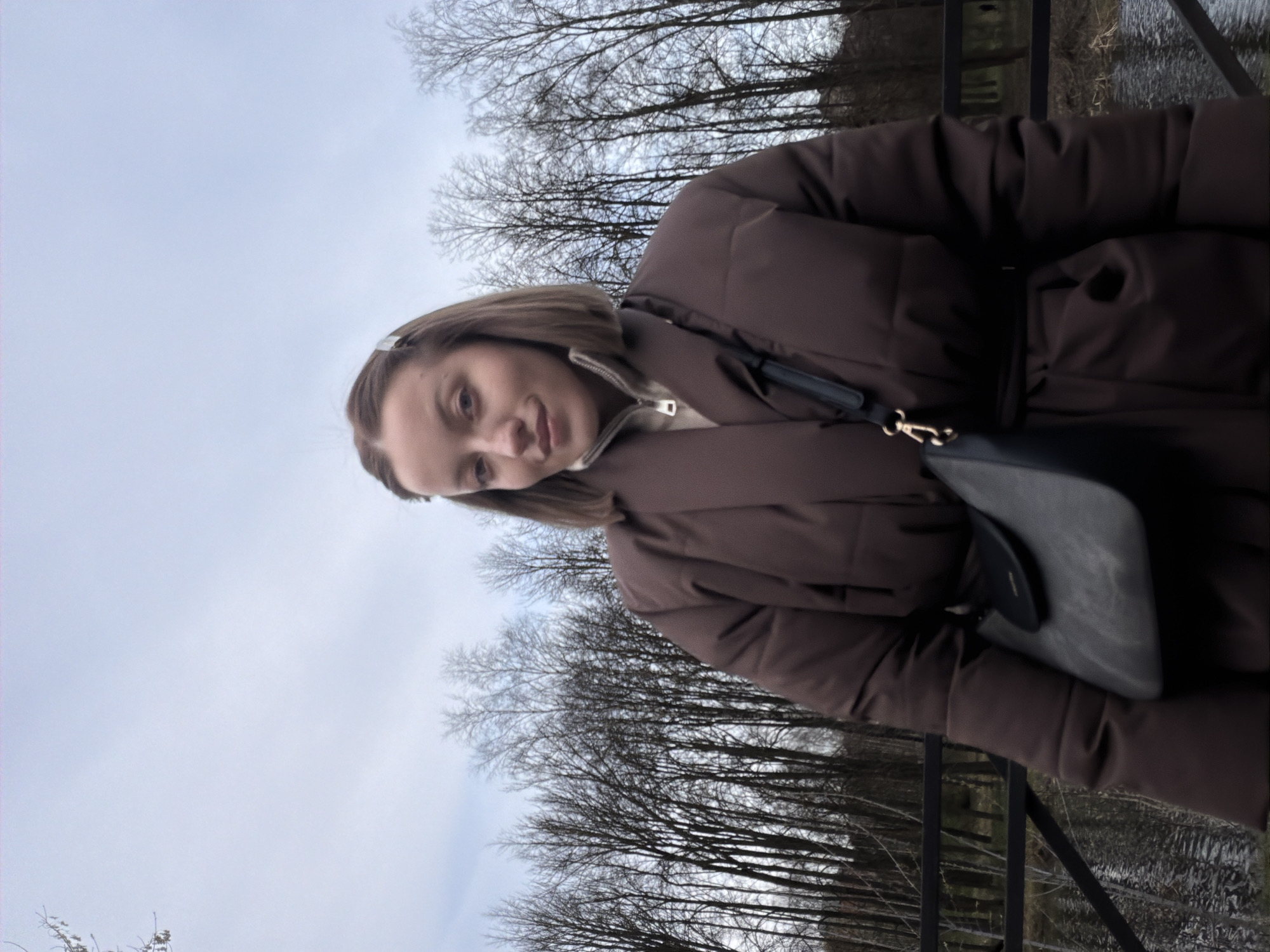 A woman stands outdoors wearing a brown puffer jacket and a light-colored top with a zipper. She is looking towards the camera. A black purse hangs from her shoulder. The background includes bare trees, a gray sky, and a metal railing. The location is Klin, Russia.
