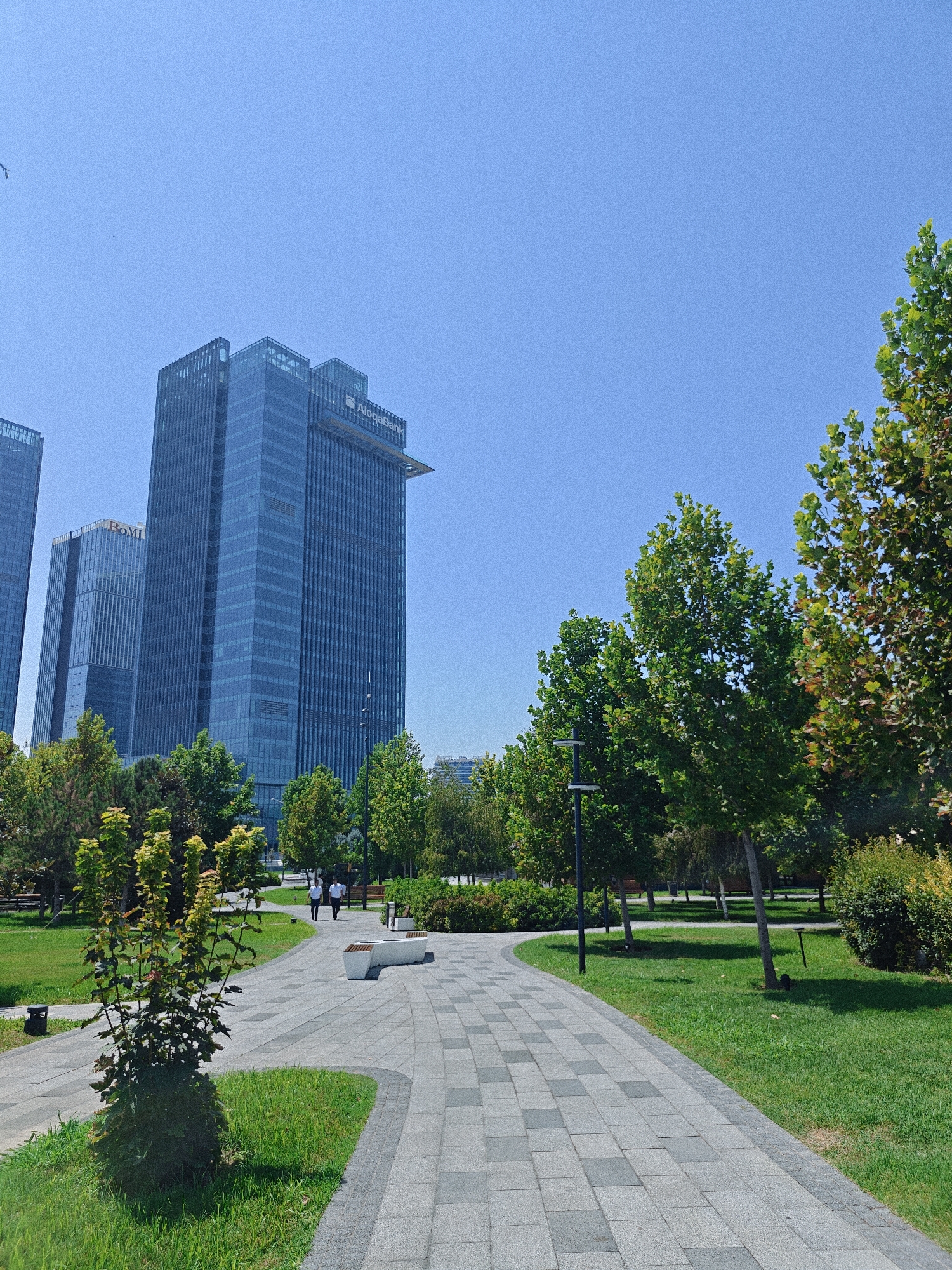 The image depicts a modern urban park in Tashkent, Uzbekistan, with tall glass skyscrapers in the background. In the foreground, a paved walkway curves through a manicured lawn dotted with trees. A few people are seen walking along the path, and a modern concrete bench is visible. The sky is clear and blue. The skyscrapers feature a distinctive architectural style with reflective glass facades. The presence of lush greenery and modern amenities suggests this is a well-maintained public space within the city.