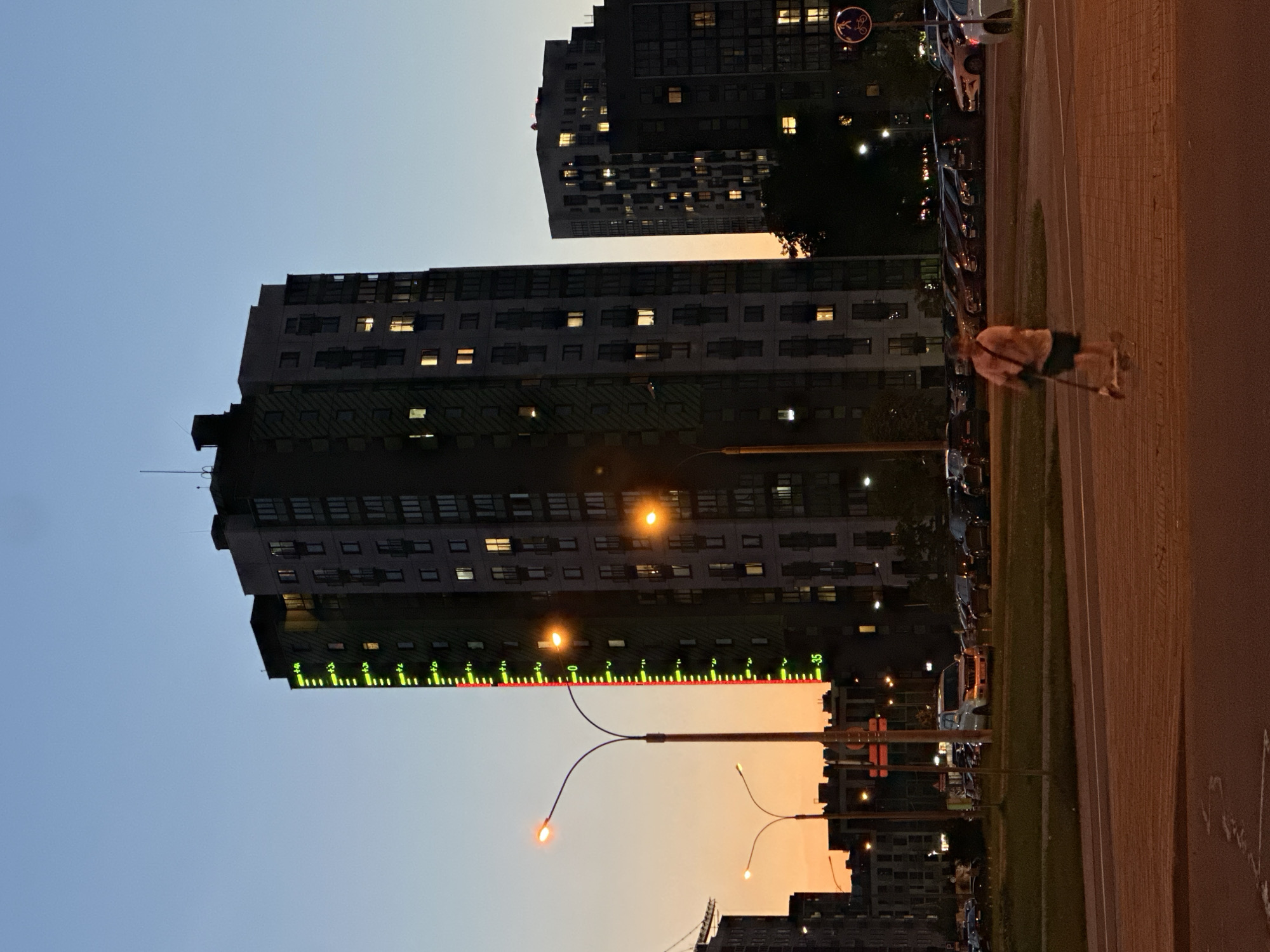 The image depicts an urban or suburban evening scene, likely set in Бараўлянскі сельскі Савет, Belarus. The sky, transitioning from a pale blue to a warm orange-yellow at the horizon, indicates the time of day is dusk or early evening.
Several multi-story apartment buildings dominate the frame. A prominent, dark-colored high-rise building occupies the left side, featuring numerous windows, many of which are illuminated, suggesting active residences. A notable detail on this building is a vertical, illuminated display running along its side, composed of green and red lights. This display shows numeric indicators such as "20," "25," "30," "35," and a red line below "-35," possibly representing a measurement scale or an artistic lighting element. Another taller, darker building is visible further back on the right, also with scattered illuminated windows.
In the foreground on the right, a wide paved road or path stretches horizontally, flanked by a grassy median strip. Beyond the median, a line of cars is parked along what appears to be a street or parking area. Several streetlights are active, emitting an orange-yellow light that contributes to the evening ambiance.
A single person is seen on the far right, walking or standing on the paved path. They are wearing a light-colored top and dark shorts and are holding a long stick, possibly a walking stick or a piece of sports equipment. A circular blue sign with a white bicycle symbol is visible above the parked cars, indicating a bicycle-friendly area. The scene conveys a tranquil, everyday evening in a residential area.