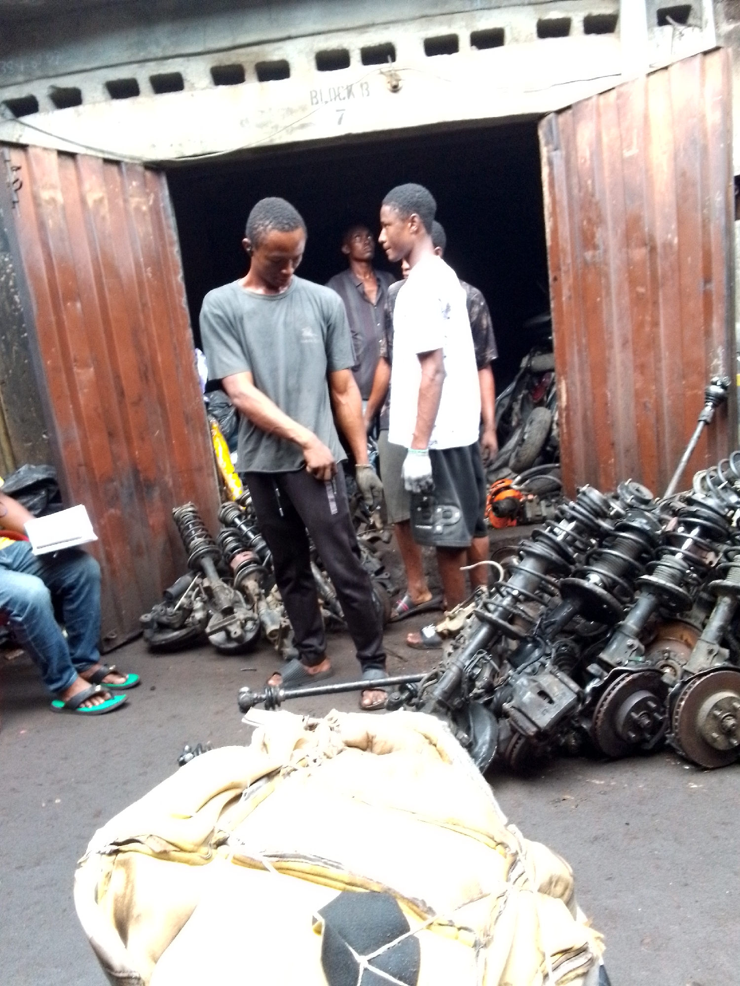 The image depicts an outdoor scene in Onitsha, Nigeria, in front of a corrugated metal structure labeled "Block 13". Several African men are present. One man, wearing a gray t-shirt and black pants, stands with his head down. Another man, wearing a white t-shirt and dark shorts, faces him. Other men are partially visible in the background, inside the metal structure. In the foreground, there is a large pile of car parts, including shock absorbers, coil springs, brake rotors, and other components. A large, white-and-black bag lies on the ground in front of the car parts. A person wearing jeans and flip-flops sits to the left, holding a notebook or papers. The overall setting appears to be an auto parts market or salvage yard.