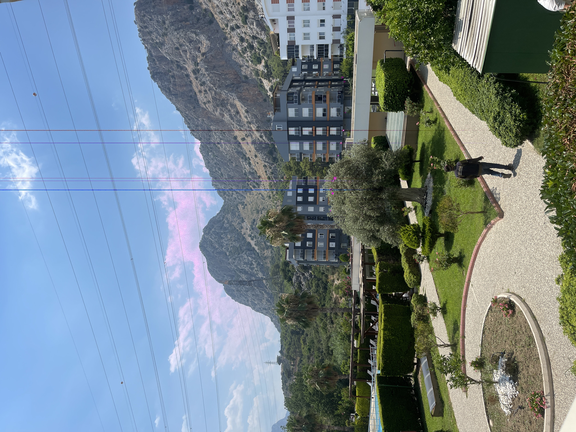 The image depicts a sunny outdoor scene in Konyaaltı, Turkey, characterized by lush greenery, modern architecture, and a mountainous backdrop. In the foreground, a well-maintained garden features a circular flower bed with white stones and pink flowers, surrounded by a winding gravel path. Adjacent to the path is a neatly trimmed hedge. Further into the scene, several trees, including a prominent olive tree with a gnarled trunk, are scattered across a grassy lawn.
Beyond the garden, a swimming pool is visible, bordered by manicured hedges and shaded seating areas with lounge chairs. Several multi-story apartment buildings with contemporary designs are situated behind the pool area. These buildings feature balconies and a mix of dark gray, white, and orange facade elements.
In the background, a large, rocky mountain dominates the landscape, with smaller hills covered in dense green vegetation leading up to it. High-voltage power lines stretch across the sky, with pylons visible on the hills. The sky is a clear blue with scattered white clouds, and some pinkish hues are present in the clouds and sky, possibly due to the lighting or camera artifact.
A single individual, a man wearing a dark jacket, blue jeans, and a beanie, is walking along the gravel path, away from the camera. His posture suggests he is moving purposefully through the grounds. The overall atmosphere is peaceful and serene, indicative of a residential or resort area.