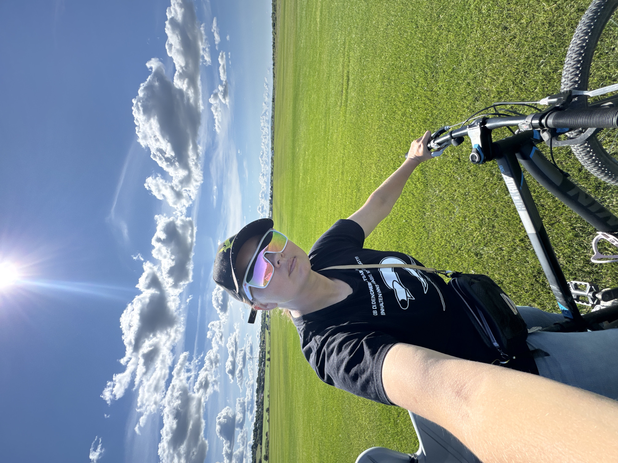 A woman wearing a black baseball cap, iridescent sunglasses, and a black t-shirt is taking a selfie with a bicycle in an open, grassy field. The sky above is a bright blue with scattered white clouds and the sun is shining brightly. The woman is holding the handlebars of the bicycle with her right hand. A small black fanny pack is worn around her waist. The bicycle appears to be a mountain bike. The background shows a vast expanse of green grass with a distant treeline and a few buildings. The overall impression is an outdoor activity on a sunny day in a rural or park-like setting, possibly on the outskirts of Kempelevo, Russia.