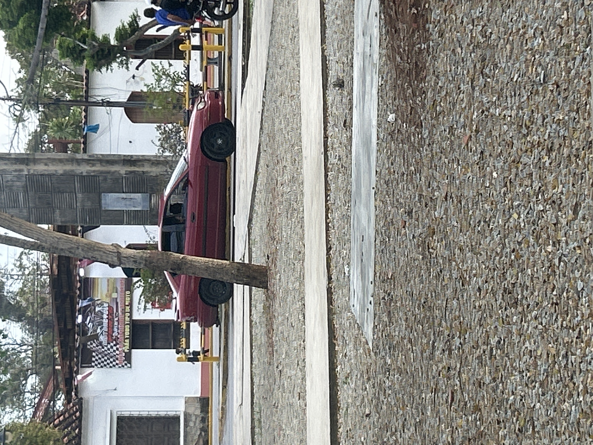 The image shows a red sedan parked on a street in Santa Ana, Venezuela. A checkered flag banner hangs from the side of a building behind the car. There are people and a motorcycle visible in the background. The street appears to be paved with cobblestones and there is a tree in the foreground.